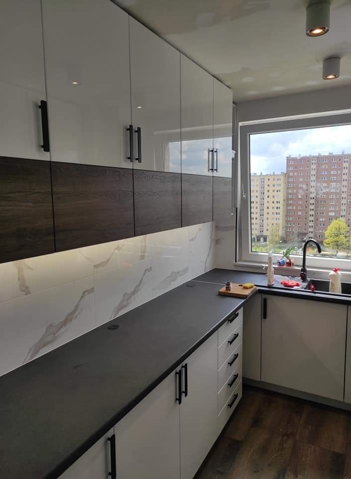 Nowoczesna kuchnia z białymi szafkami z czarnymi uchwytami, ciemnym blatem i marmurowymi płytkami nad blatem, widok na blok mieszkalny za oknem.