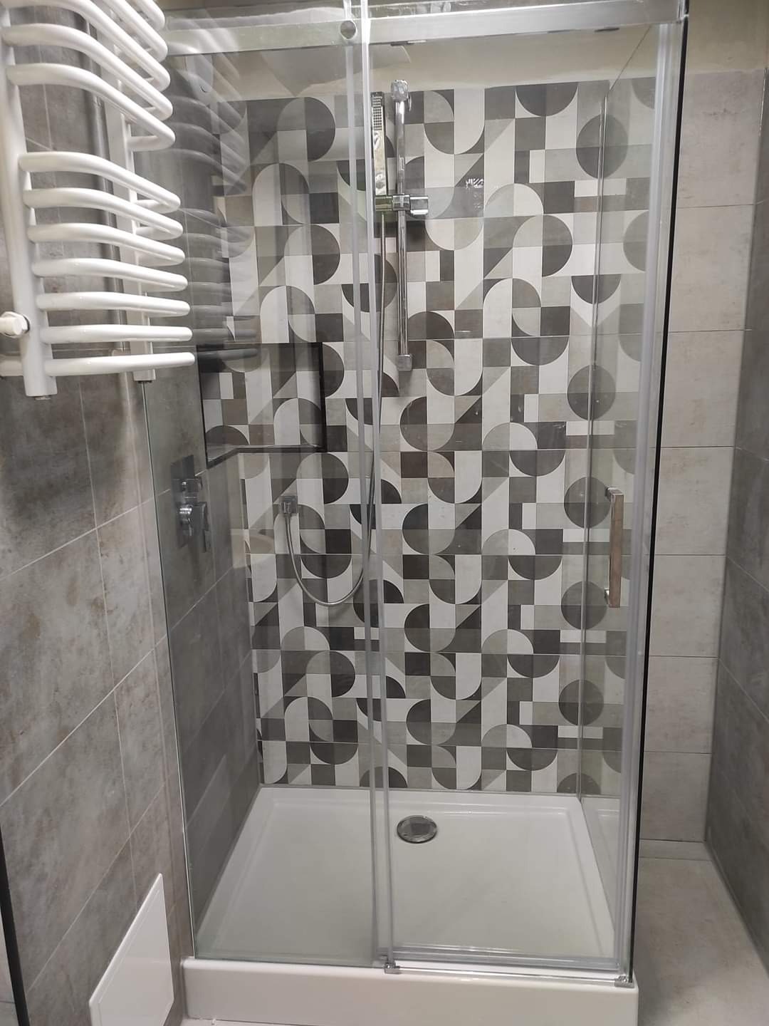 Nowoczesna kabina prysznicowa z przesuwnymi drzwiami, biały brodzik, ściana wyłożona płytkami w geometryczne wzory w odcieniach szarości, biały grzejnik drabinkowy po lewej stronie.