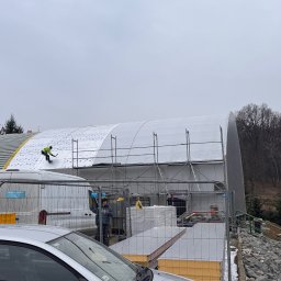 Grzegorz Tomasiak GT-BAU - Pracownik w żółtej kamizelce montuje białe płyty na dachu hali przemysłowej, widoczny fragment rusztowania, samochody dostawcze i częściowo samochód osobowy na pierwszym planie, pochmurne niebo w tle.