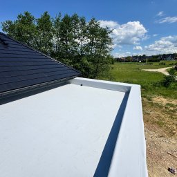 Grzegorz Tomasiak GT-BAU - Widok z góry na biały, płaski dach z widocznymi obróbkami blacharskimi, fragment dachu pokrytego czarną dachówką oraz zielone otoczenie w tle.