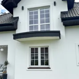 Fragment białego domu z ciemną dach&oacute;wką, rynnami i stolarką okienną. Widoczne okna z białymi szprosami i czarne lampy elewacyjne.