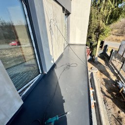 Grzegorz Tomasiak GT-BAU - Wykończony, czarny taras z widocznym narzędziem (opalarka) i oknem z odbiciem czerwonego samochodu. Widok z góry na wykończoną powierzchnię i otoczenie budynku.