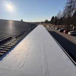 Dach budynku z częściowo ukończonym kryciem: ciemna dachówka ceramiczna kontrastuje z jasną, nową membraną dachową. W tle parking i drzewa w słoneczny dzień.