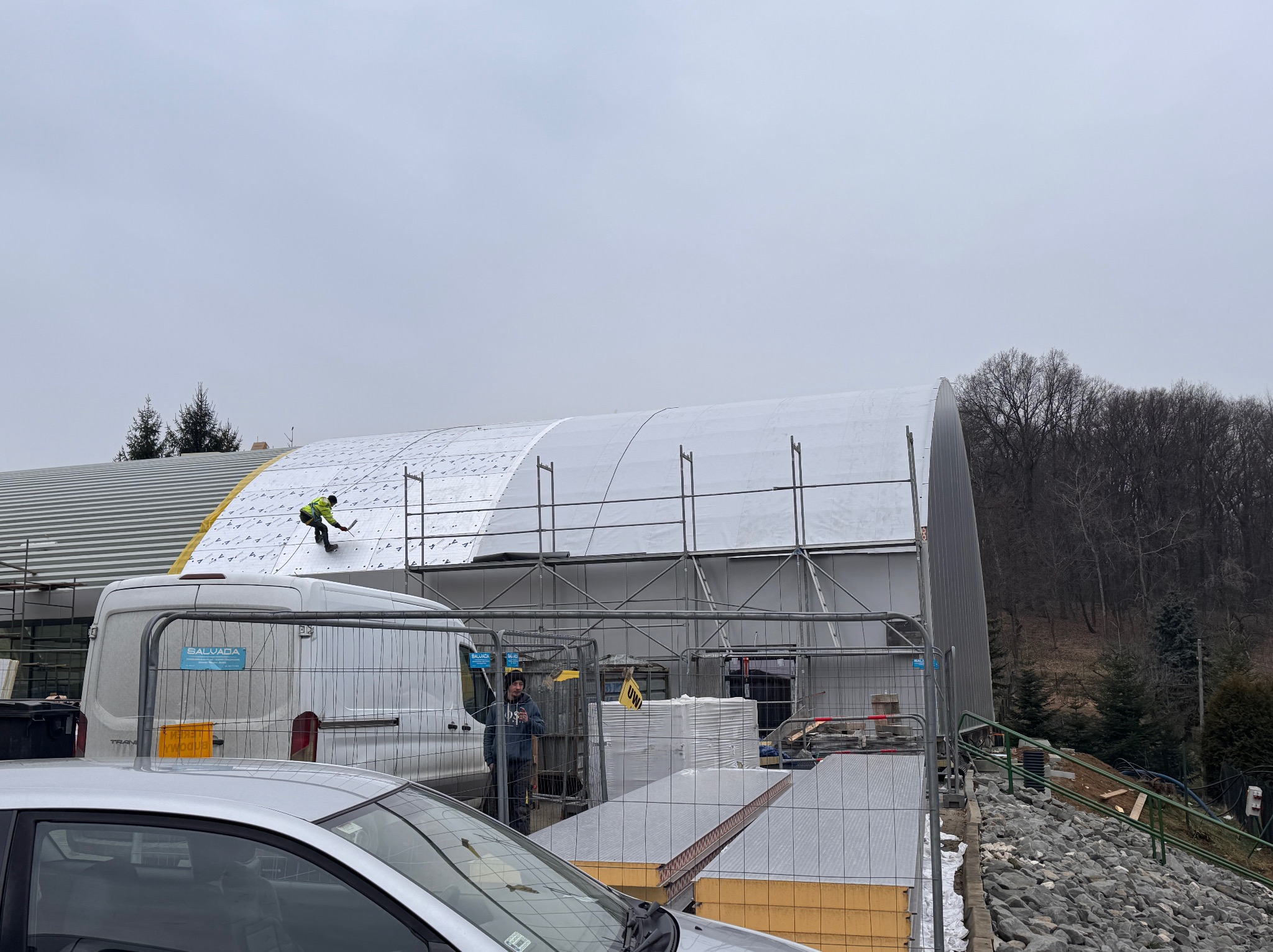 Pracownik w żółtej kamizelce montuje białe płyty na dachu hali przemysłowej, widoczny fragment rusztowania, samochody dostawcze i częściowo samochód osobowy na pierwszym planie, pochmurne niebo w tle.