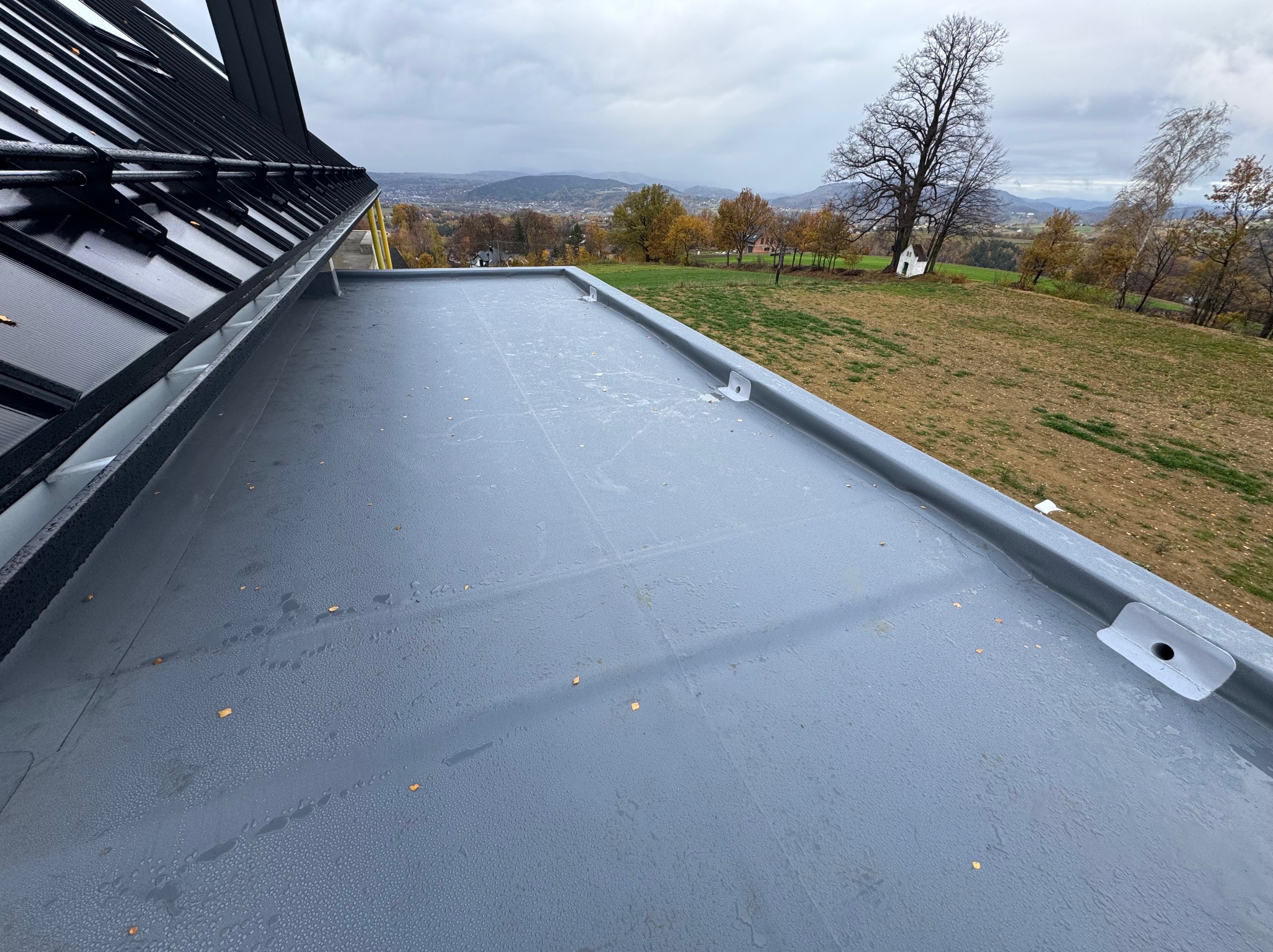 Szary, płaski dach z widocznymi mocowaniami i rynną, z jesiennym krajobrazem w tle pod pochmurnym niebem.