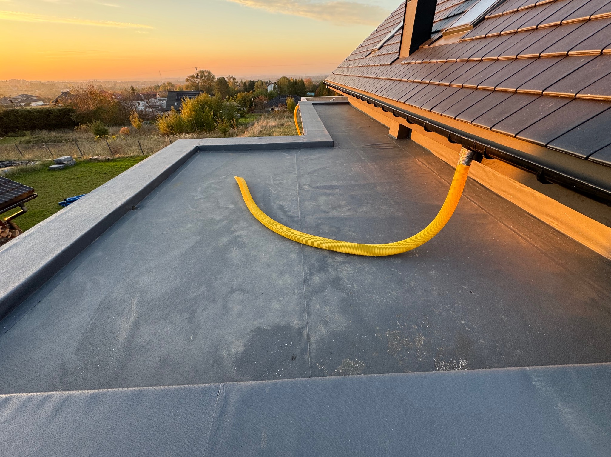 Płaski dach pokryty czarną membraną bitumiczną z żółtym wężem odprowadzającym wodę do rynny. W tle widok na zabudowania i zachodzące słońce.