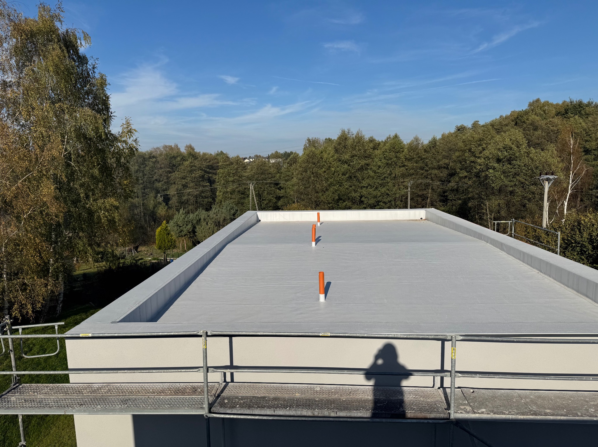 Płaski dach pokryty szarą membraną hydroizolacyjną z trzema pomarańczowymi odpowietrznikami, widok z rusztowania na tle lasu i błękitnego nieba.
