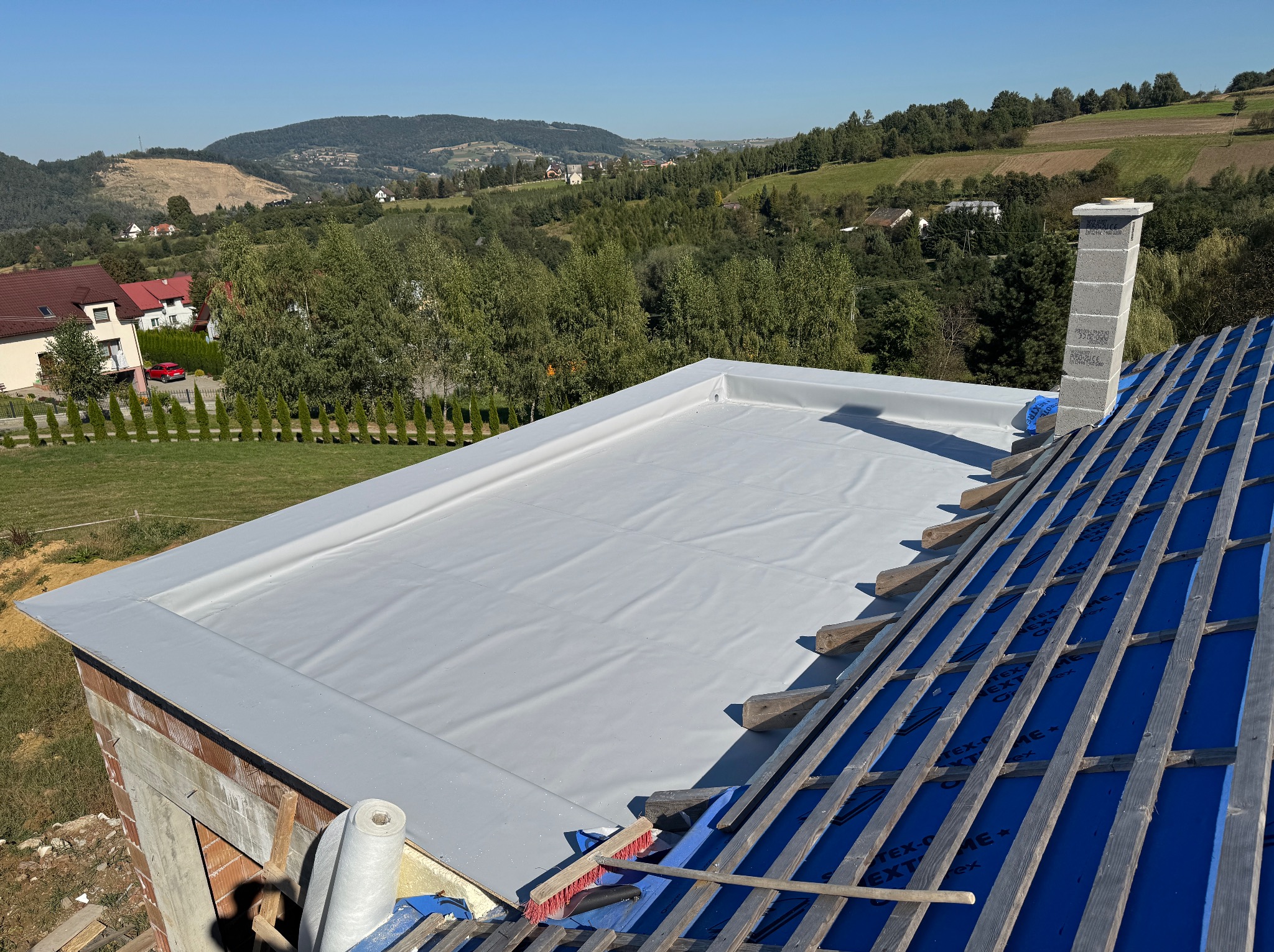 Wykończony płaski dach pokryty szarą membraną PVC, widoczne krokwie z niebieską folią w tle, komin z bloczków betonowych i malowniczy krajobraz zalesionych wzgórz.