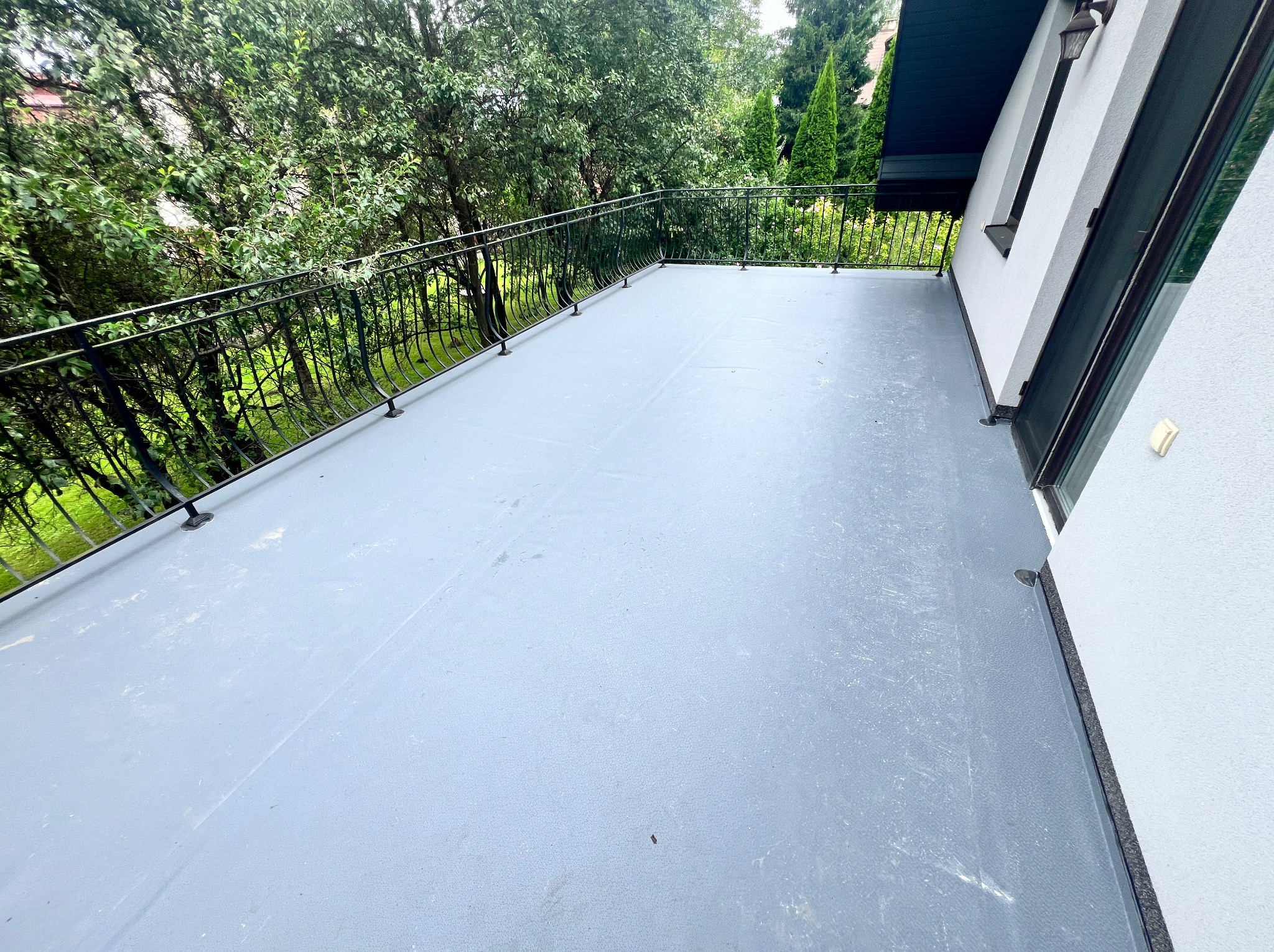 Szary balkon z metalową balustradą, widok na zieleń, nowoczesne wykończenie elewacji budynku.