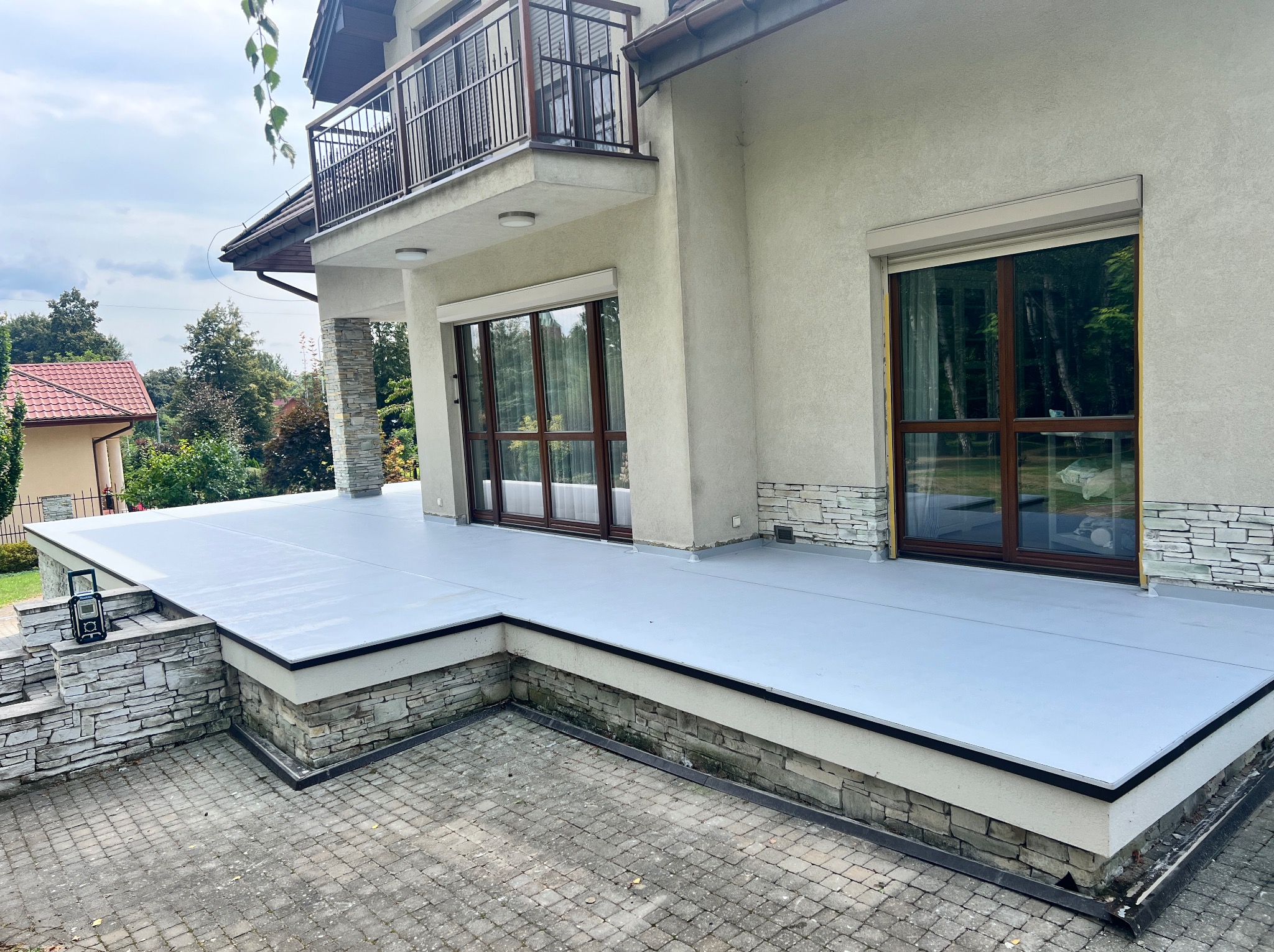 Nowoczesny taras z szarą, płaską powierzchnią, wykończony kamieniem elewacyjnym, widok z poziomu kostki brukowej, okna z brązowymi ramami w tle.