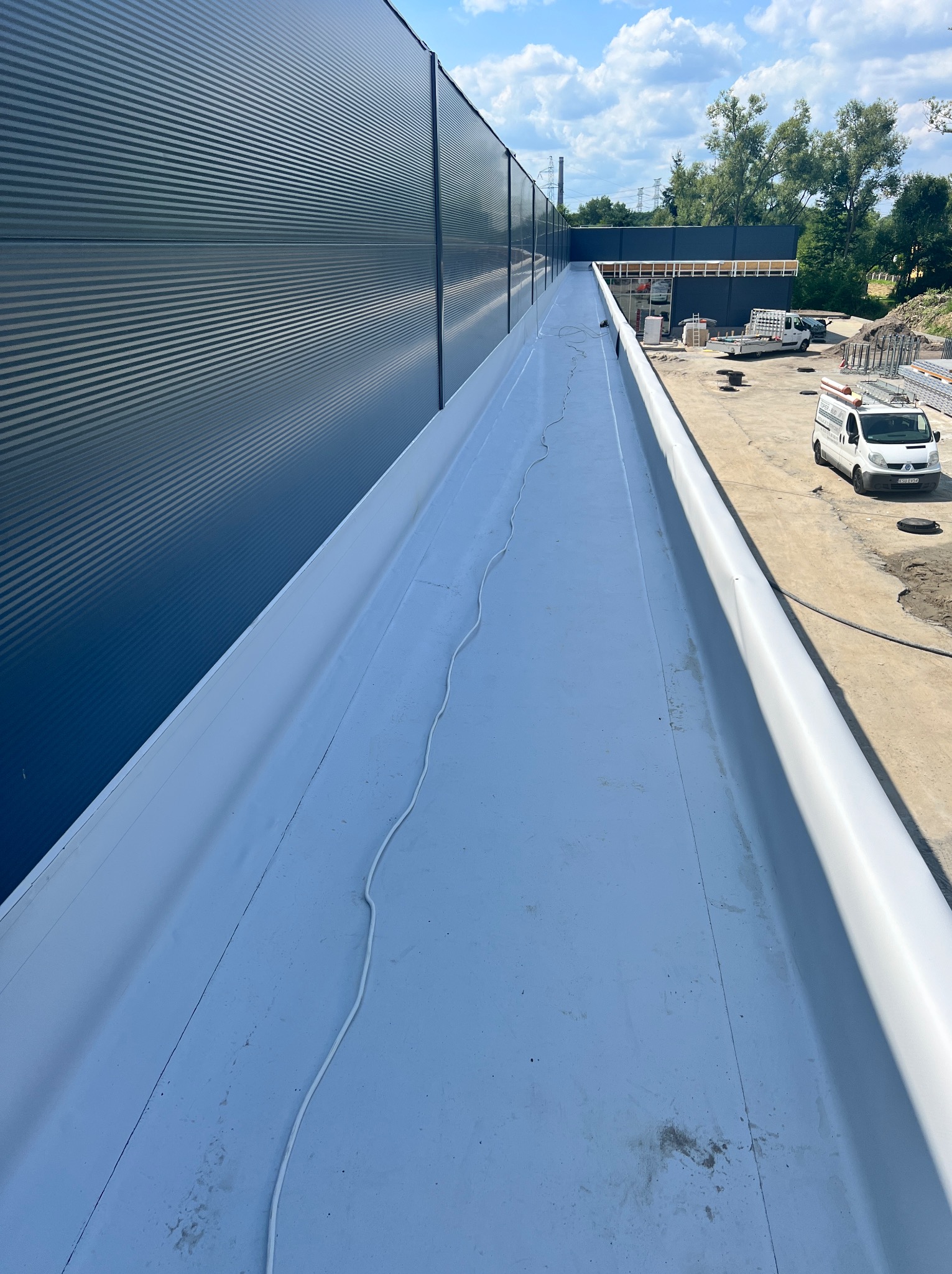 Płaski dach pokryty białą membraną, widoczny przewód elektryczny, fragment ściany z blachy trapezowej, w tle zaparkowany biały samochód dostawczy i materiały budowlane.