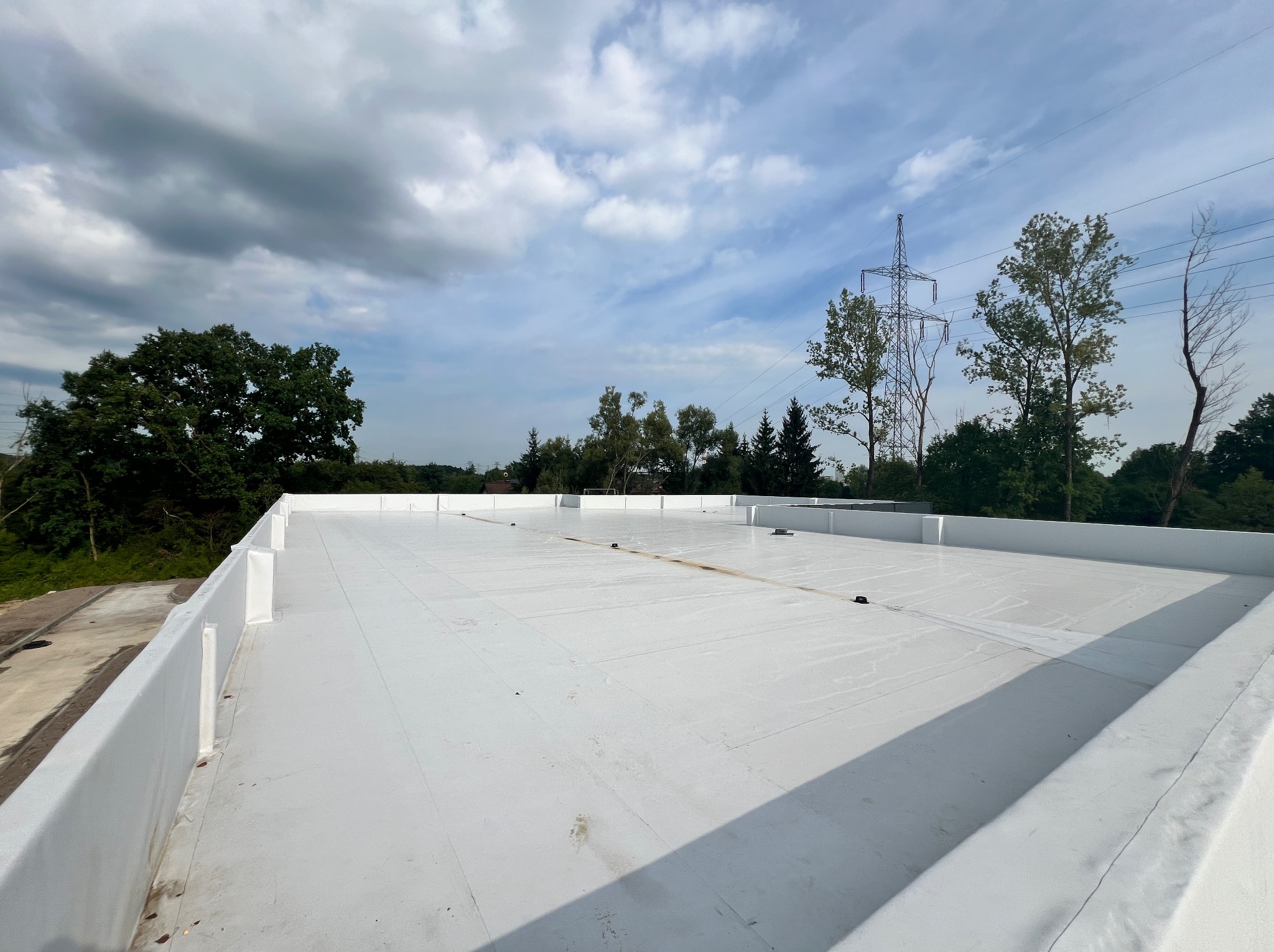 Biały, płaski dach z widocznymi łączeniami, otoczony niską ścianką attykową, na tle pochmurnego nieba i zieleni drzew oraz słupa wysokiego napięcia.