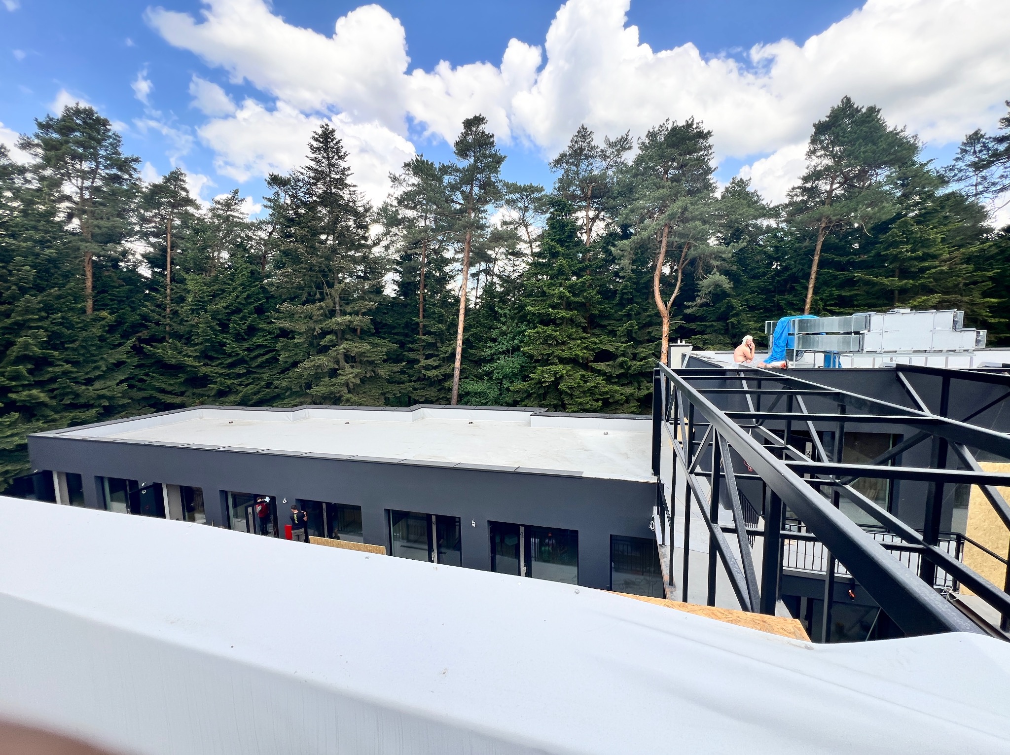 Widok z góry na płaski dach budynku z pracownikami i elementami konstrukcyjnymi, w tle las iglasty i błękitne niebo z chmurami.