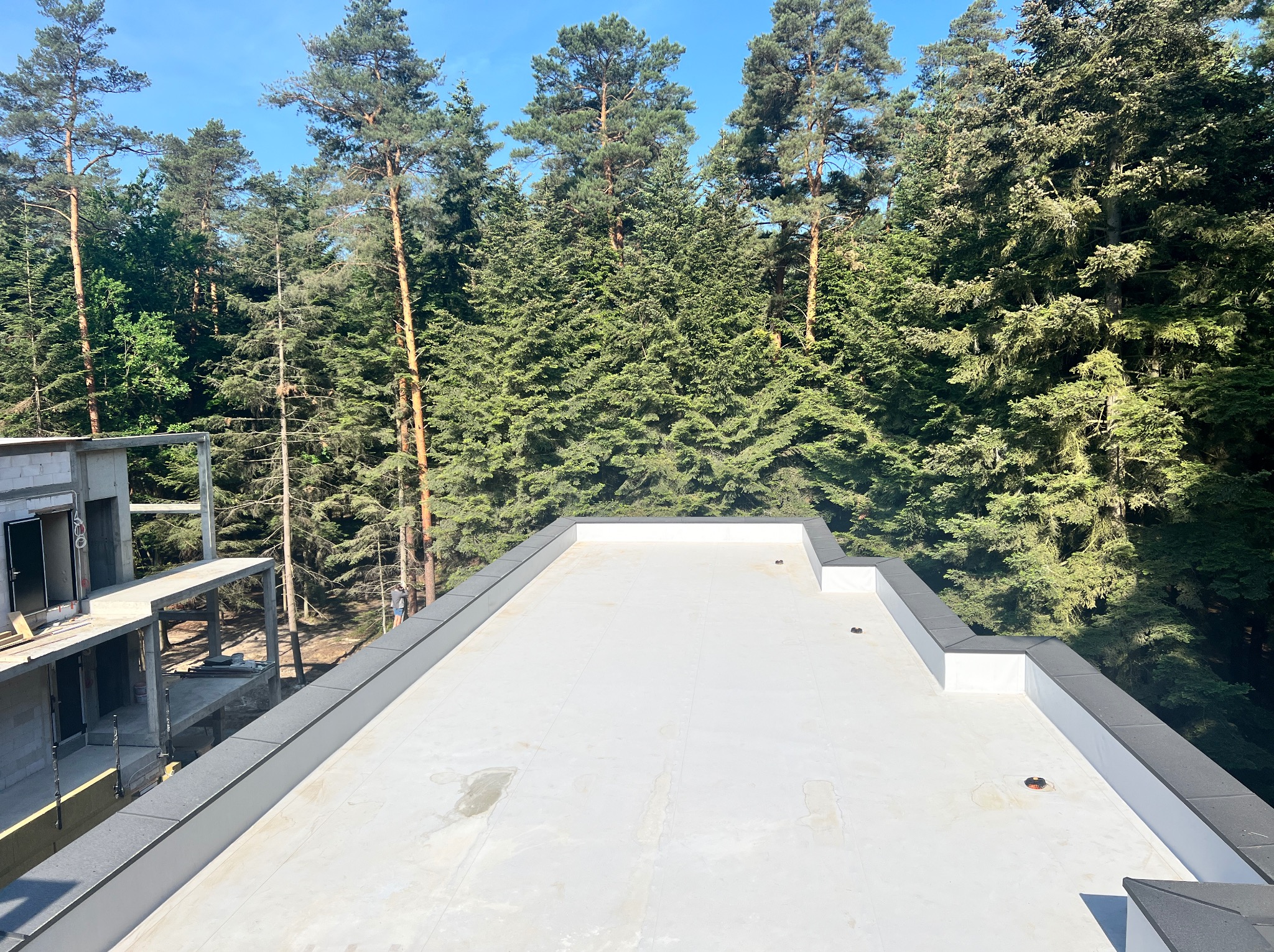 Biały, płaski dach nowego budynku w stanie surowym, otoczony zielenią drzew, widok z góry.