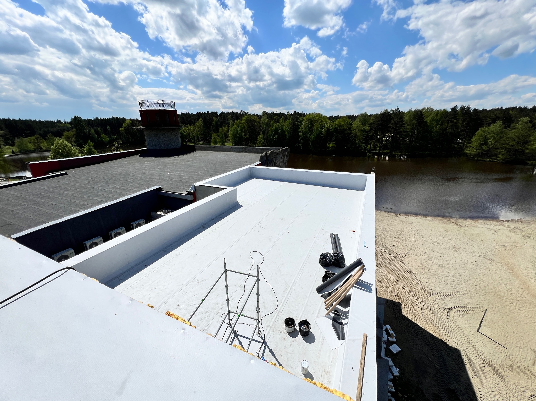 Widok z góry na fragment dachu płaskiego z nową białą membraną, częściowo wykończonego, z widocznym rusztowaniem, materiałami budowlanymi i elementami wentylacji, w tle las i zbiornik wodny.
