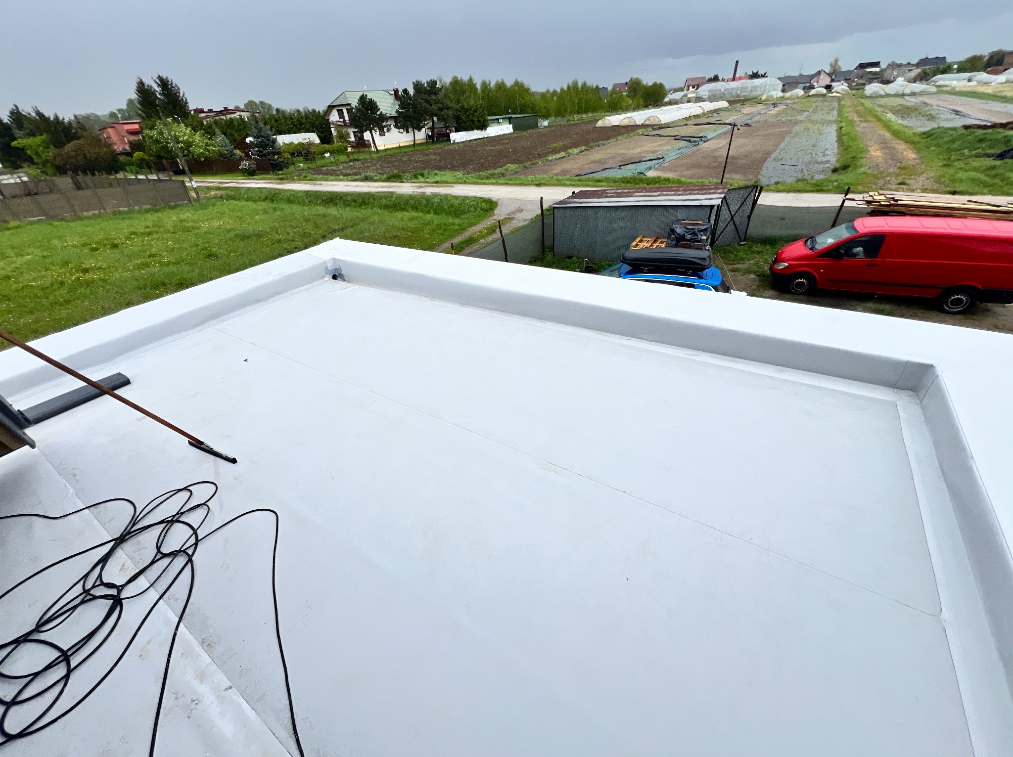 Biały, płaski dach z widocznymi liniami łączeń membrany, na tle zabudowań gospodarczych, pól uprawnych i pochmurnego nieba; widoczne narzędzia i przewód elektryczny.