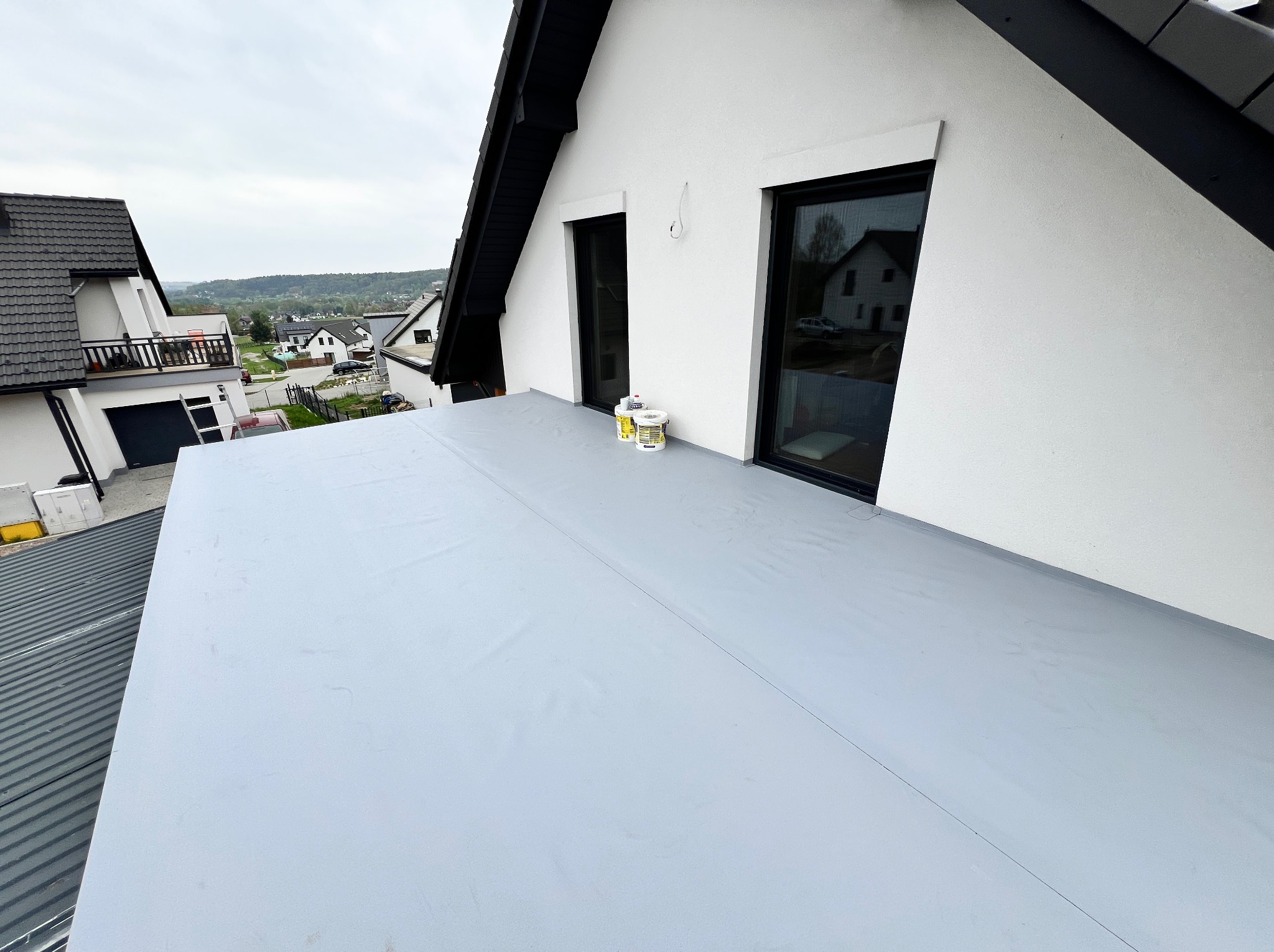 Szary, płaski dach pokryty membraną, widoczne dwa okna dachowe, puszki z materiałami budowlanymi, w tle zabudowa jednorodzinna.