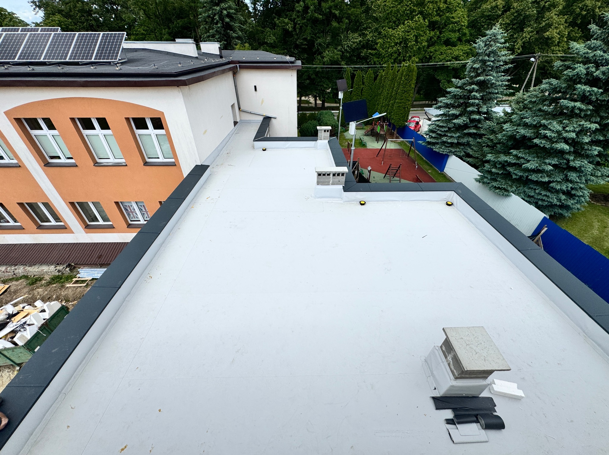Biały, płaski dach po renowacji z widocznymi elementami wykończeniowymi, kominem i panelami słonecznymi na sąsiednim dachu. W tle zieleń drzew i plac zabaw.