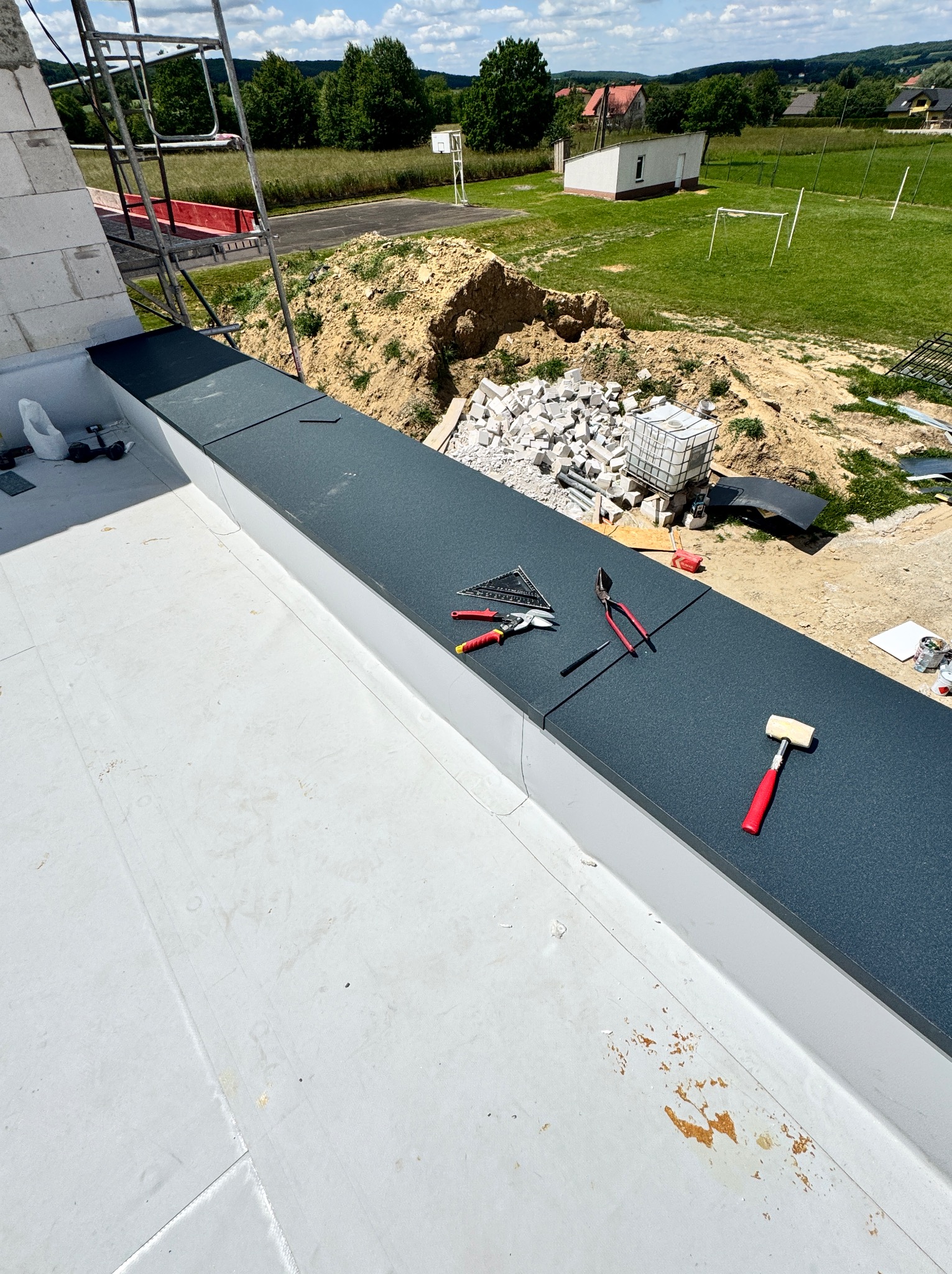 Czarny panel na dachu z narzędziami dekarskimi: młotek, kombinerki, kątownik. W tle budowa i zieleń. Widok z góry na fragment dachu w trakcie prac.