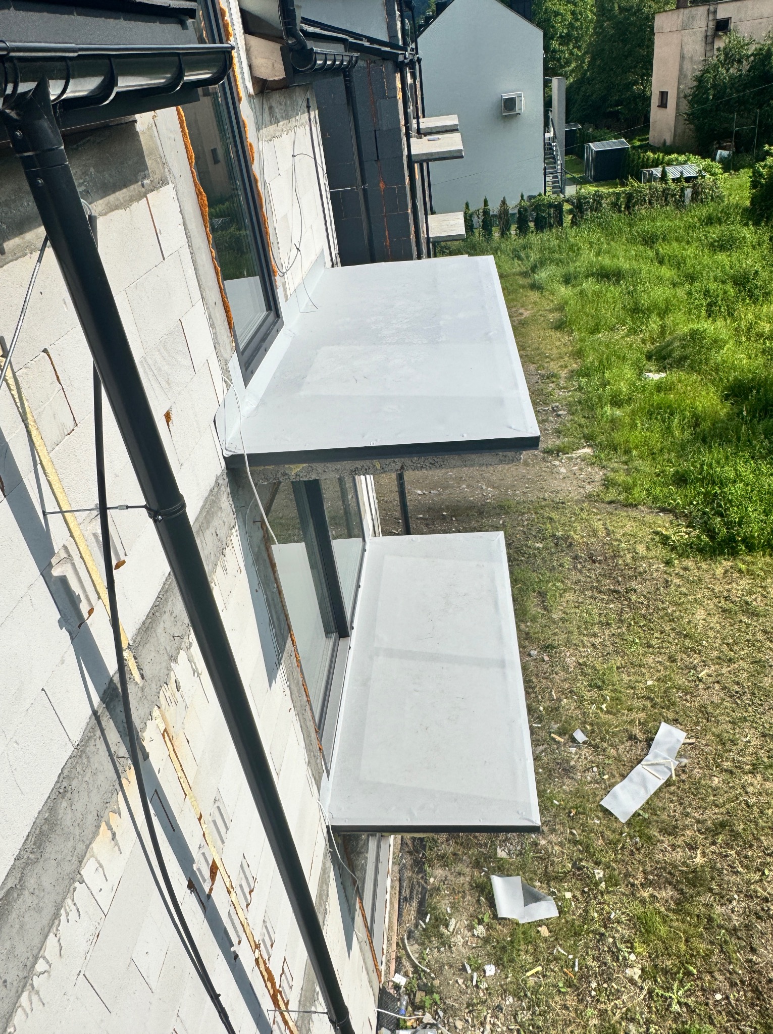 Nowe, płaskie balkony z szarą hydroizolacją, zamontowane na surowej fasadzie budynku z widocznymi rynnami spustowymi i elementami wykończeniowymi na trawie.