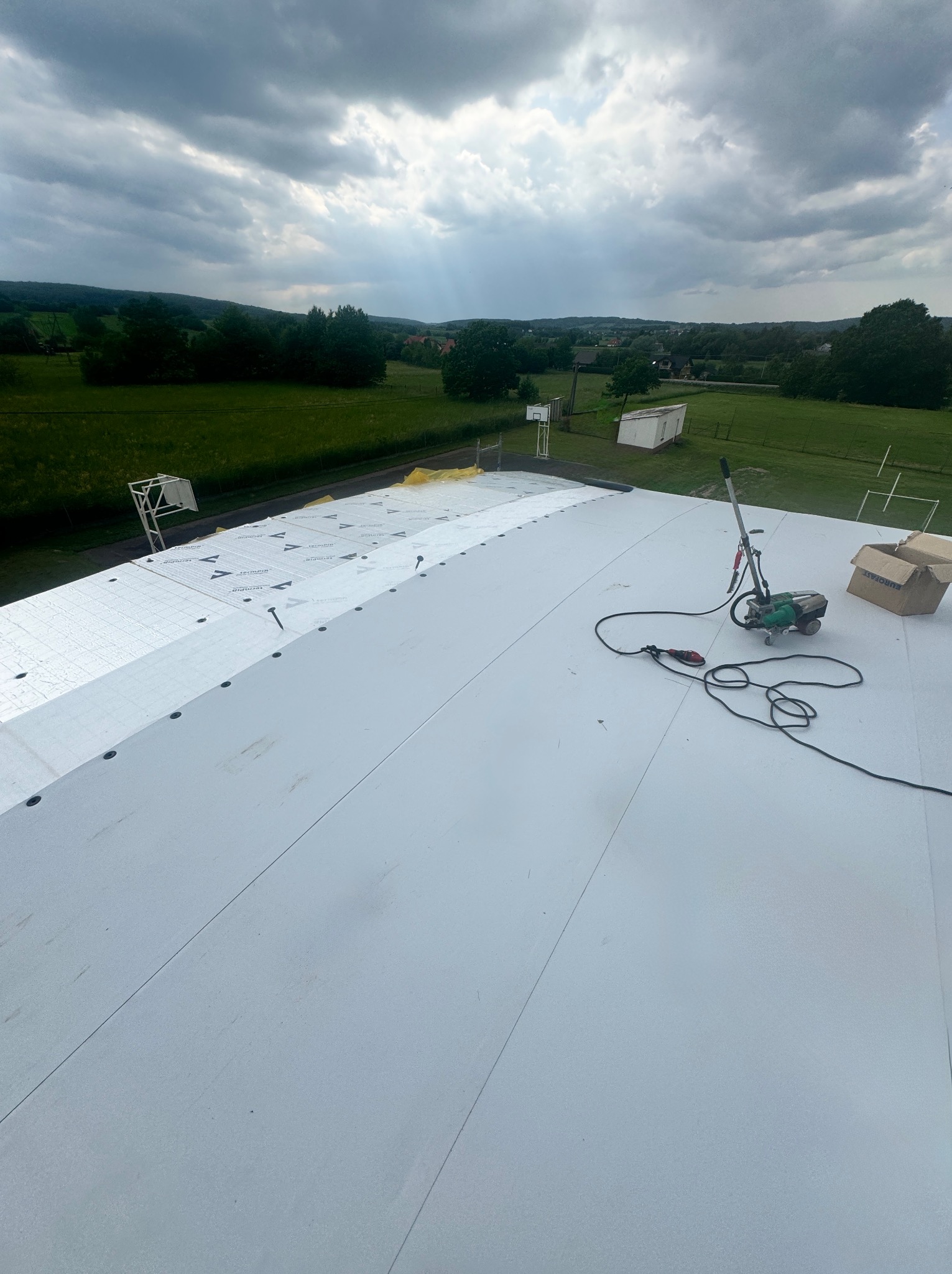 Biały dach z termoizolacją w trakcie prac wykończeniowych, widok z góry na narzędzia i krajobraz w tle, pochmurne niebo.