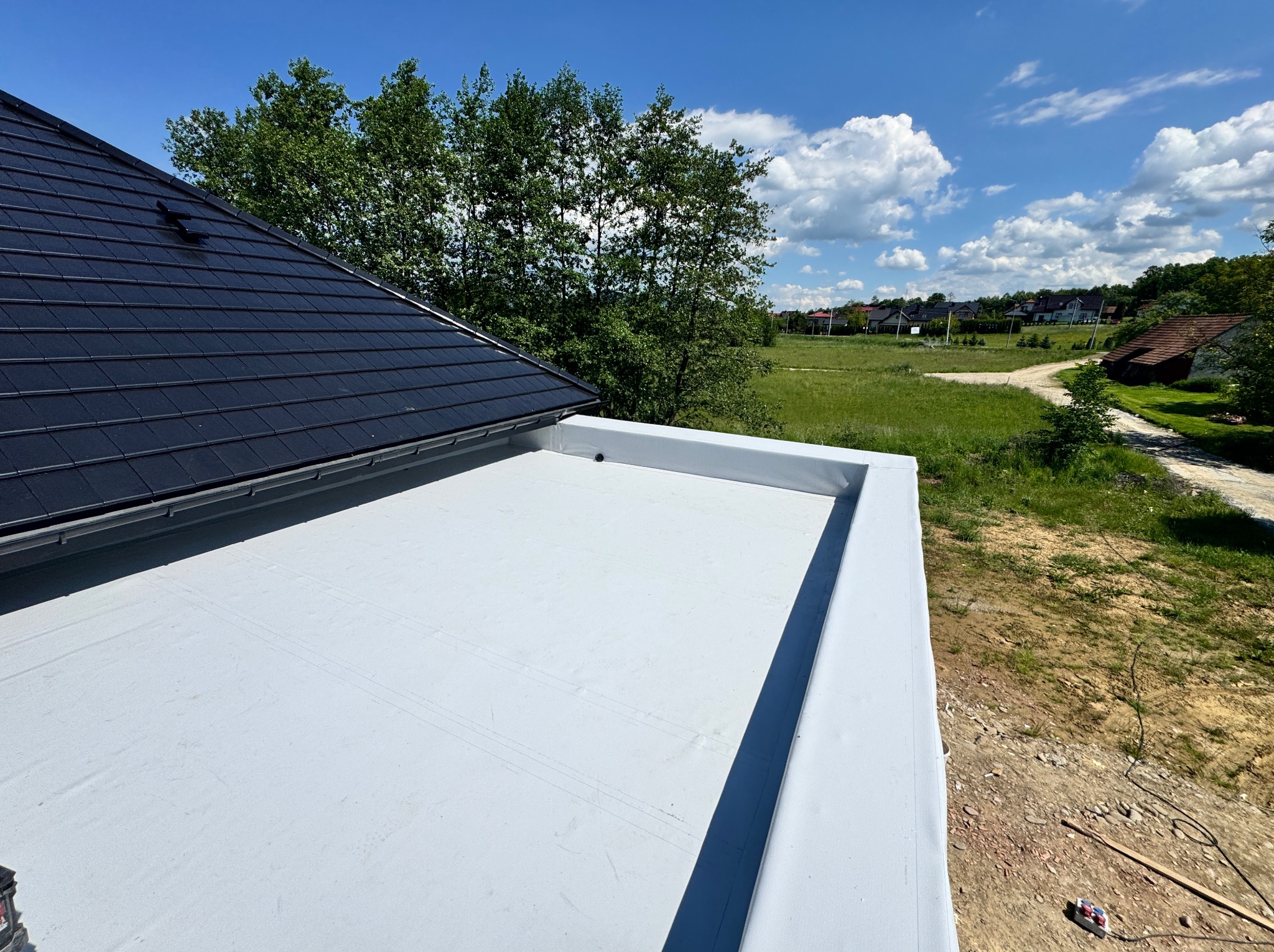 Widok z góry na biały, płaski dach z widocznymi obróbkami blacharskimi, fragment dachu pokrytego czarną dachówką oraz zielone otoczenie w tle.