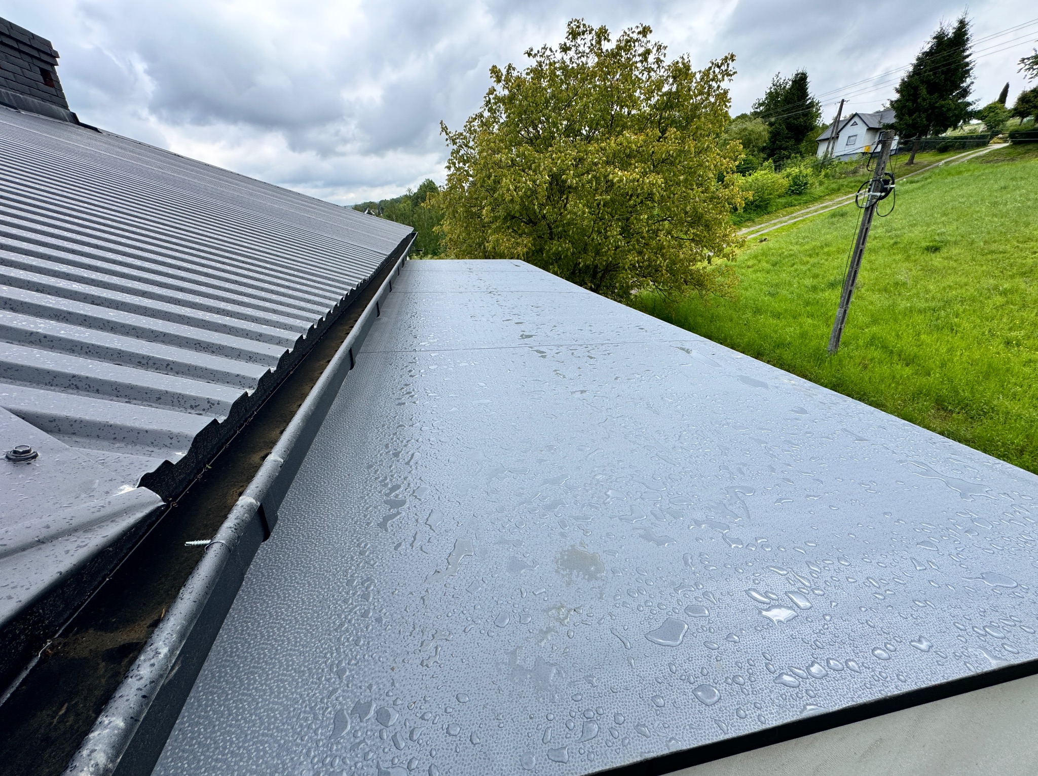 Widok z góry na nowy, szary dach z blachy trapezowej z zamontowaną rynną, pokryty kroplami deszczu; w tle zieleń drzew i trawy, pochmurne niebo.