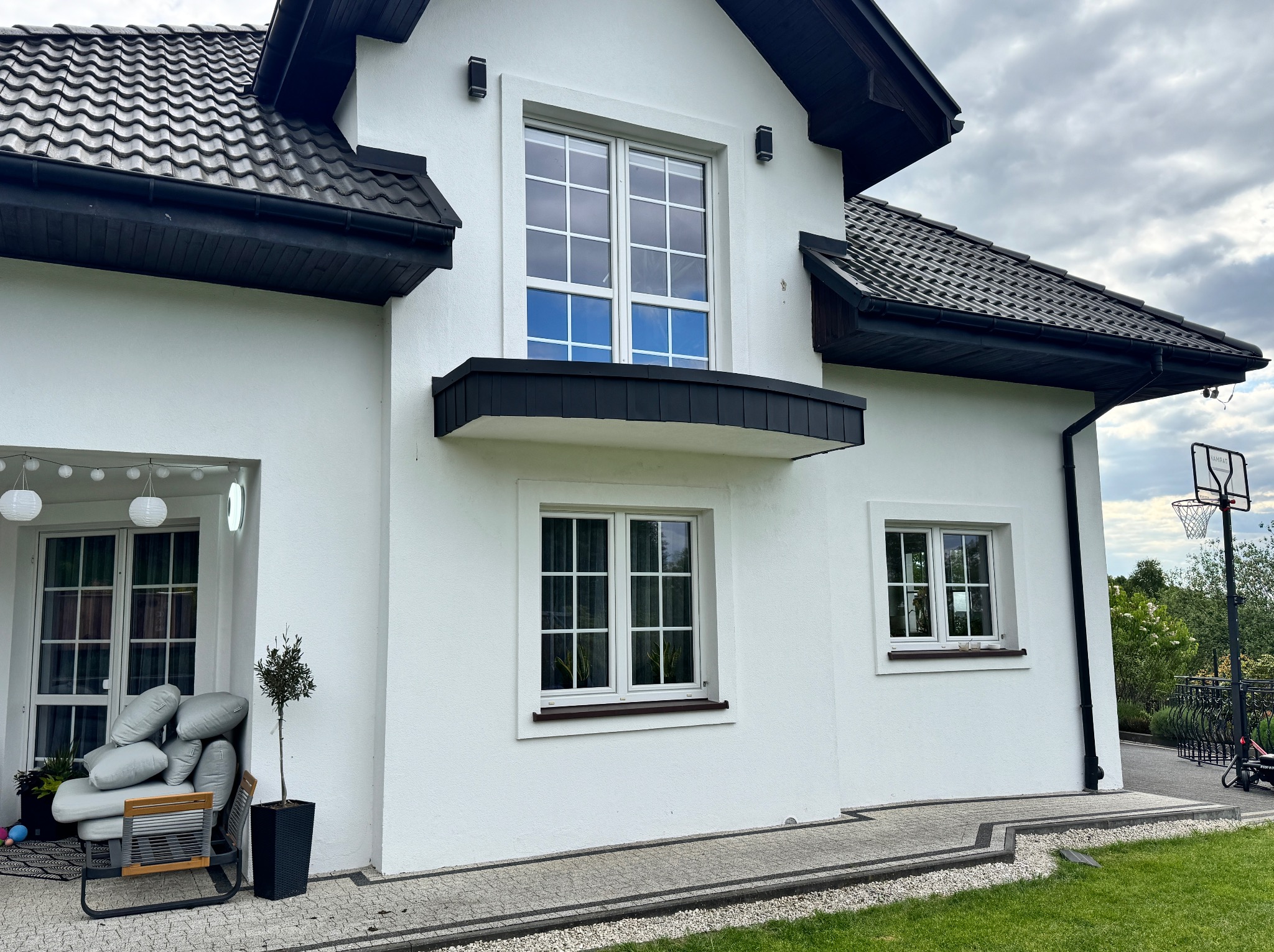 Nowoczesny dom z ciemną dachówką ceramiczną, białymi ścianami i dopasowanymi rynnami. Widoczny fragment elewacji i ogrodu z elementami architektury.