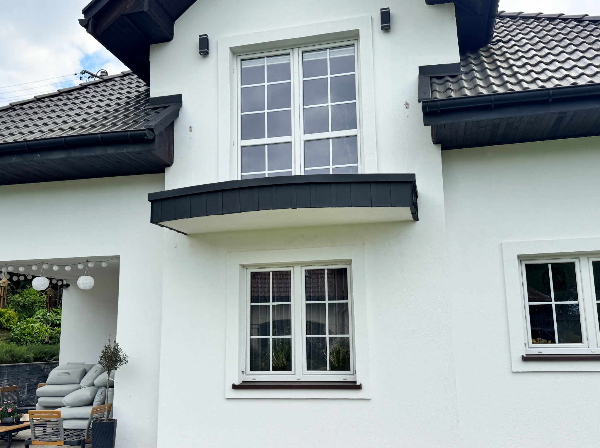 Fragment białego domu z ciemną dachówką, rynnami i stolarką okienną. Widoczne okna z białymi szprosami i czarne lampy elewacyjne.