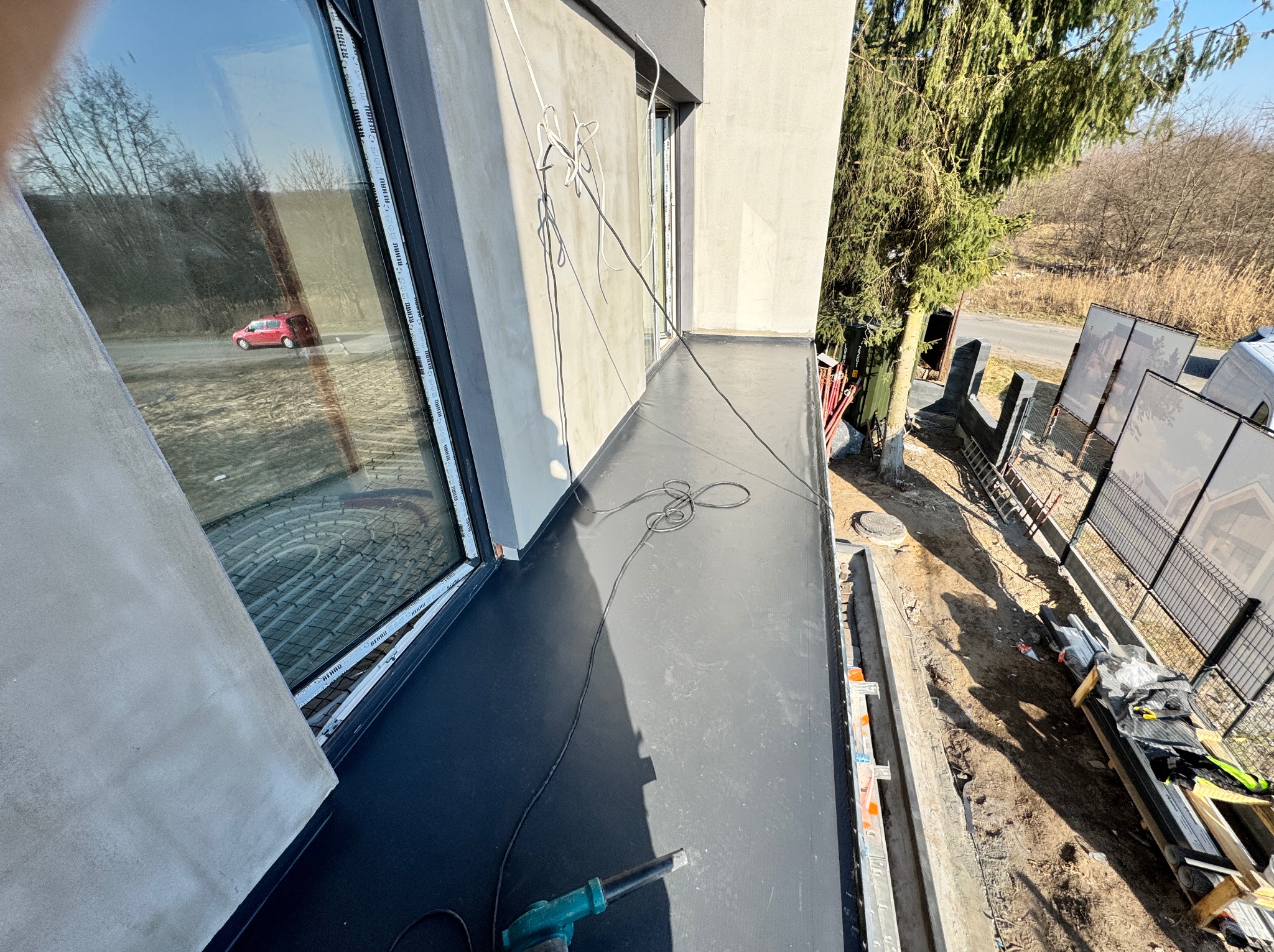 Wykończony, czarny taras z widocznym narzędziem (opalarka) i oknem z odbiciem czerwonego samochodu. Widok z góry na wykończoną powierzchnię i otoczenie budynku.