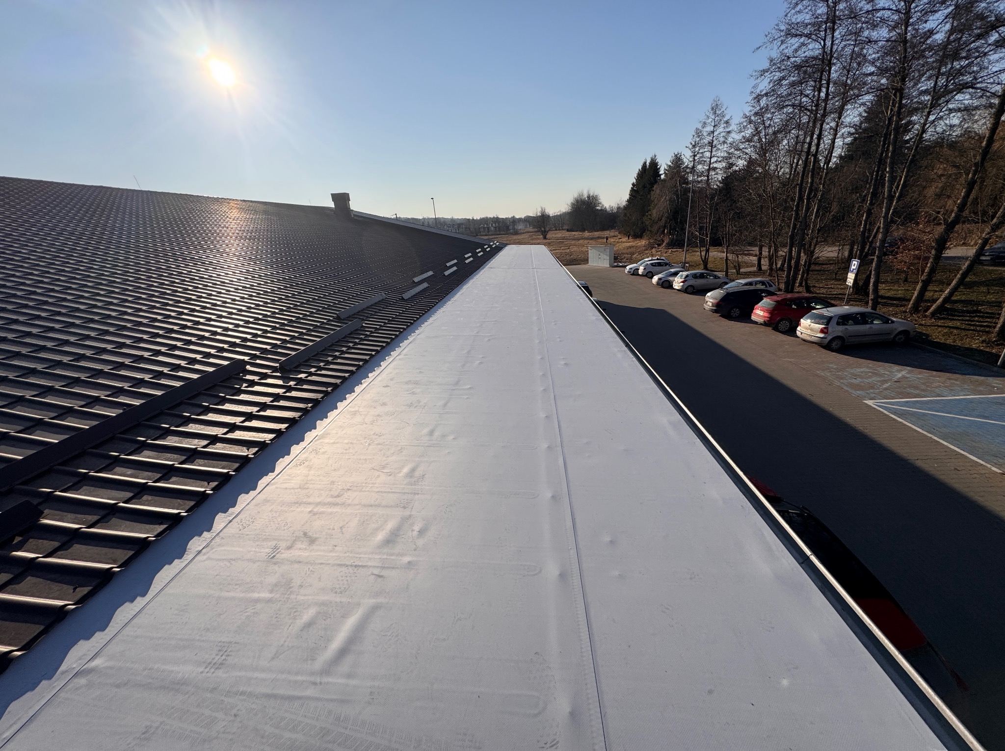 Dach budynku z częściowo ukończonym kryciem: ciemna dachówka ceramiczna kontrastuje z jasną, nową membraną dachową. W tle parking i drzewa w słoneczny dzień.