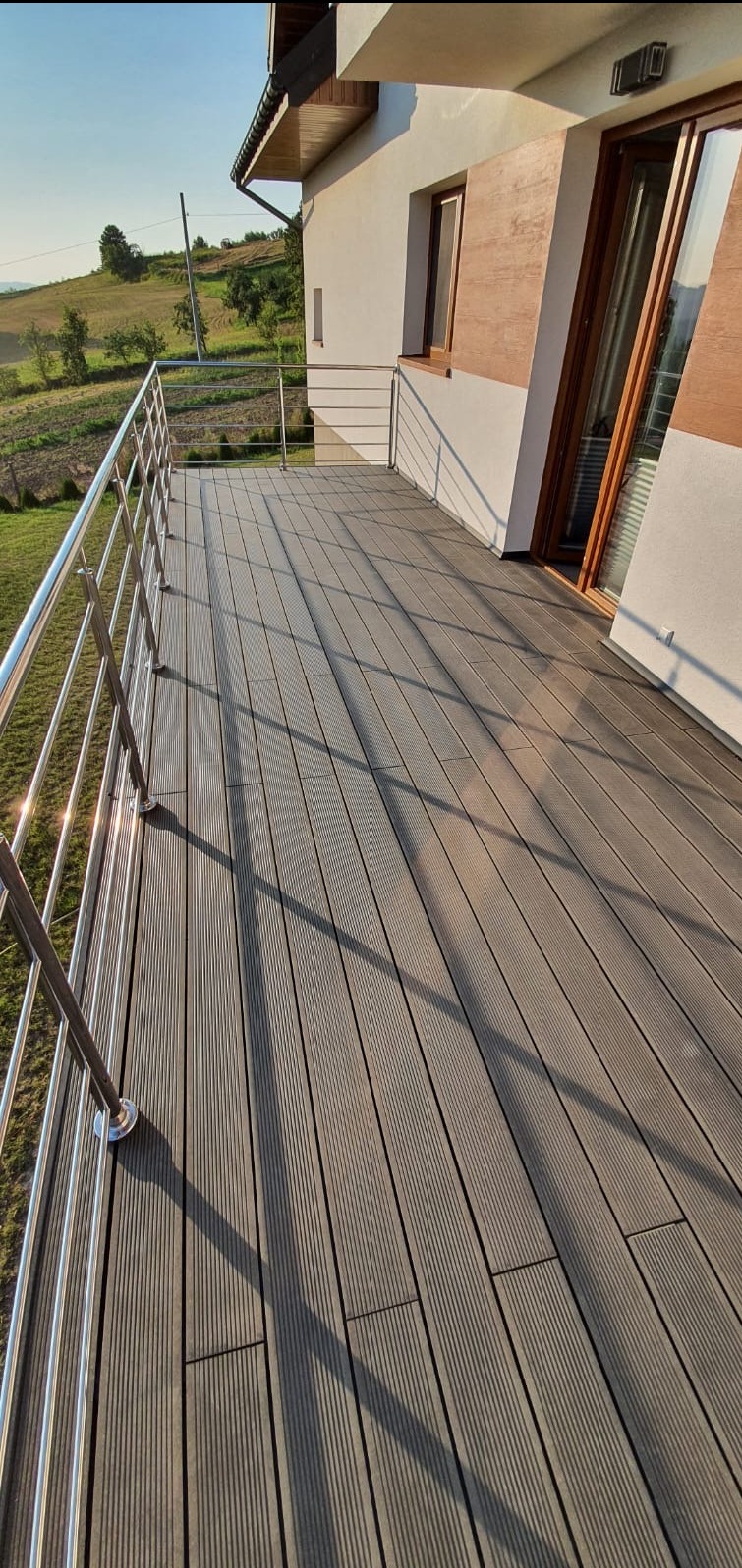 Nowoczesny balkon z szarą deską kompozytową i metalową balustradą, widok na zielone wzgórza w tle. Słoneczne oświetlenie podkreśla fakturę desek.