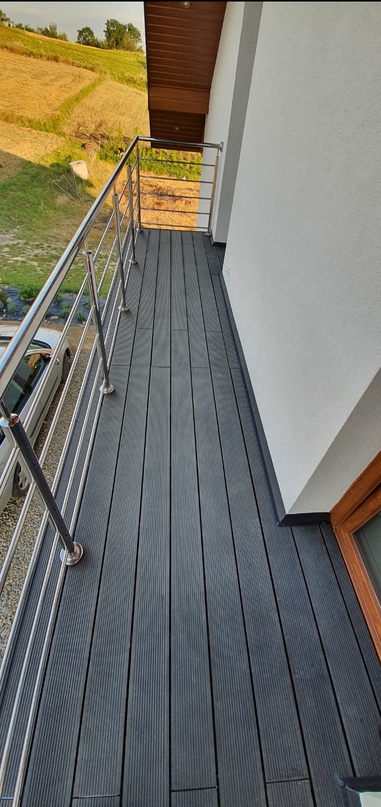 Nowoczesny balkon z szarą deską kompozytową i metalową balustradą. Widok na zielone wzgórza w tle. Estetyczne wykończenie, detale architektoniczne.