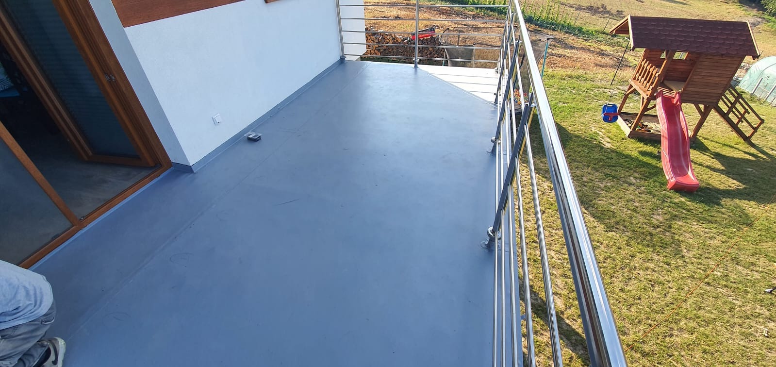 Balkon po remoncie z szarą, gładką podłogą i metalową balustradą. W tle plac zabaw i trawnik. Widok z góry, prezentujący efekt hydroizolacji i wykończenia.