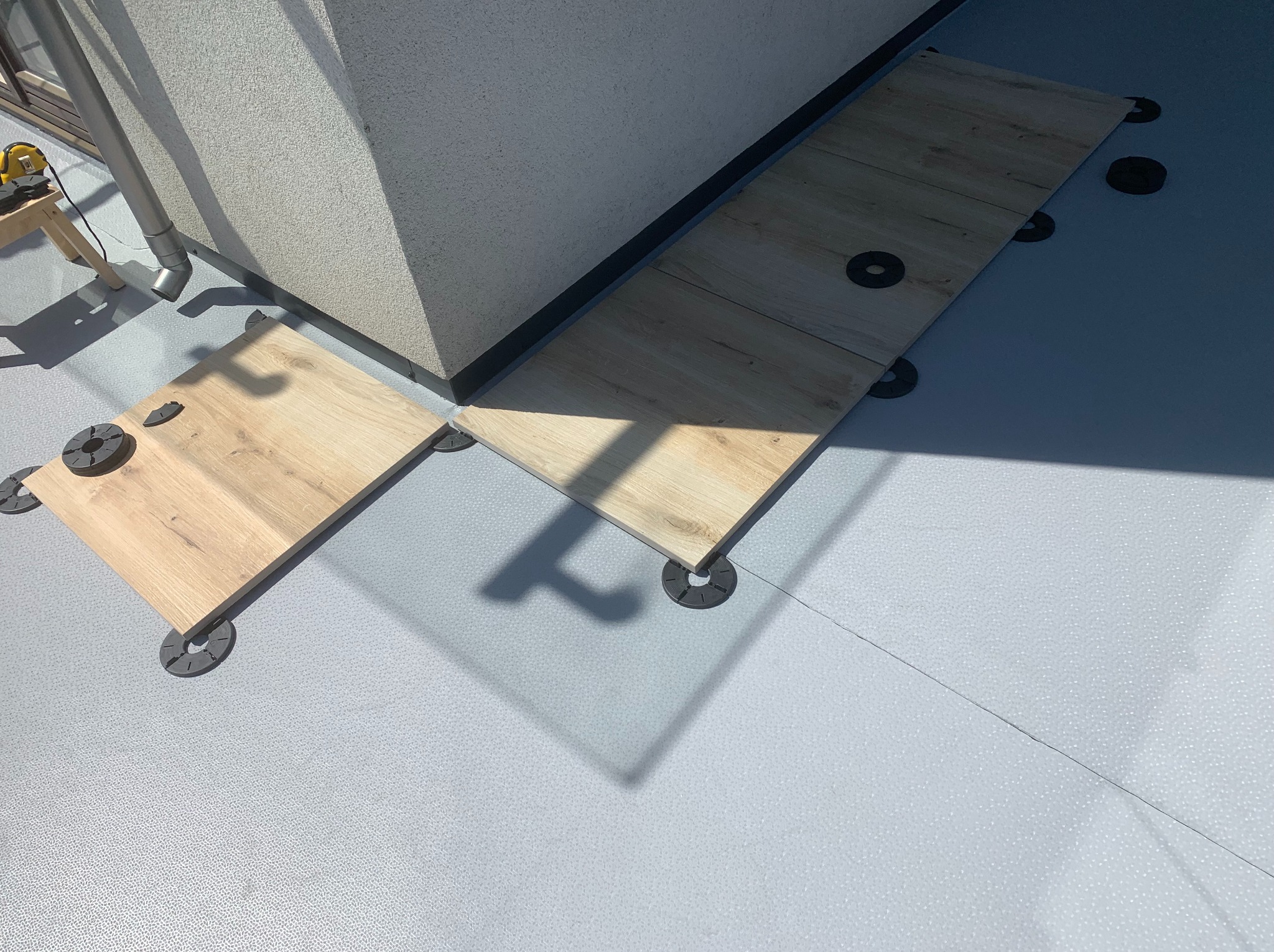 Układanie płytek na wspornikach na balkonie. Widoczne płytki imitujące drewno i szara mata izolacyjna. Częściowo ukończony montaż.