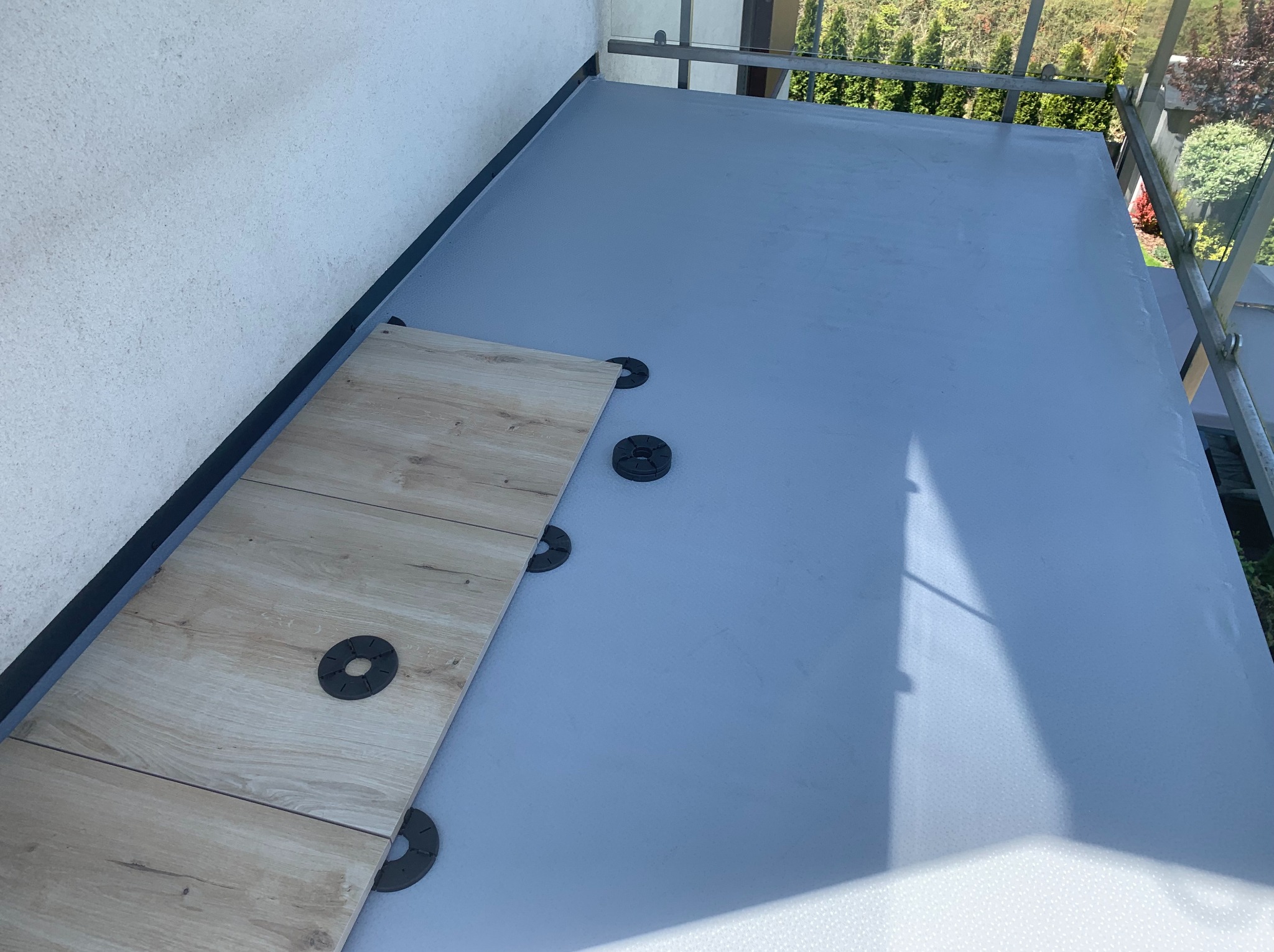 Balkon w trakcie układania płytek na specjalnych podkładkach dystansowych, widoczna hydroizolacja. Częściowo ułożone płytki w odcieniach beżu.