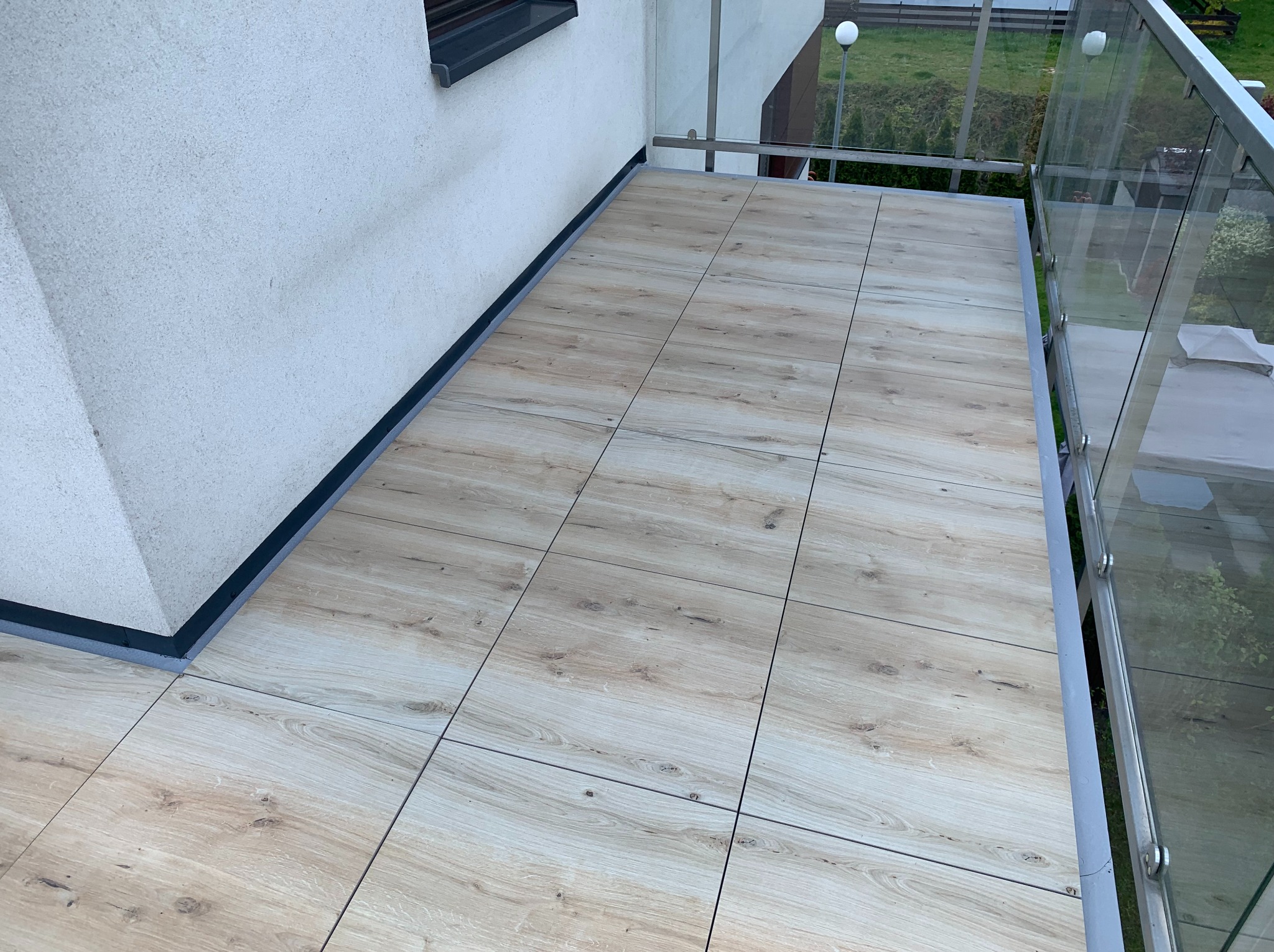 Wykończony balkon z jasnymi płytkami imitującymi drewno, szklane balustrady i widok na zieleń. Nowoczesny design i dbałość o detale.
