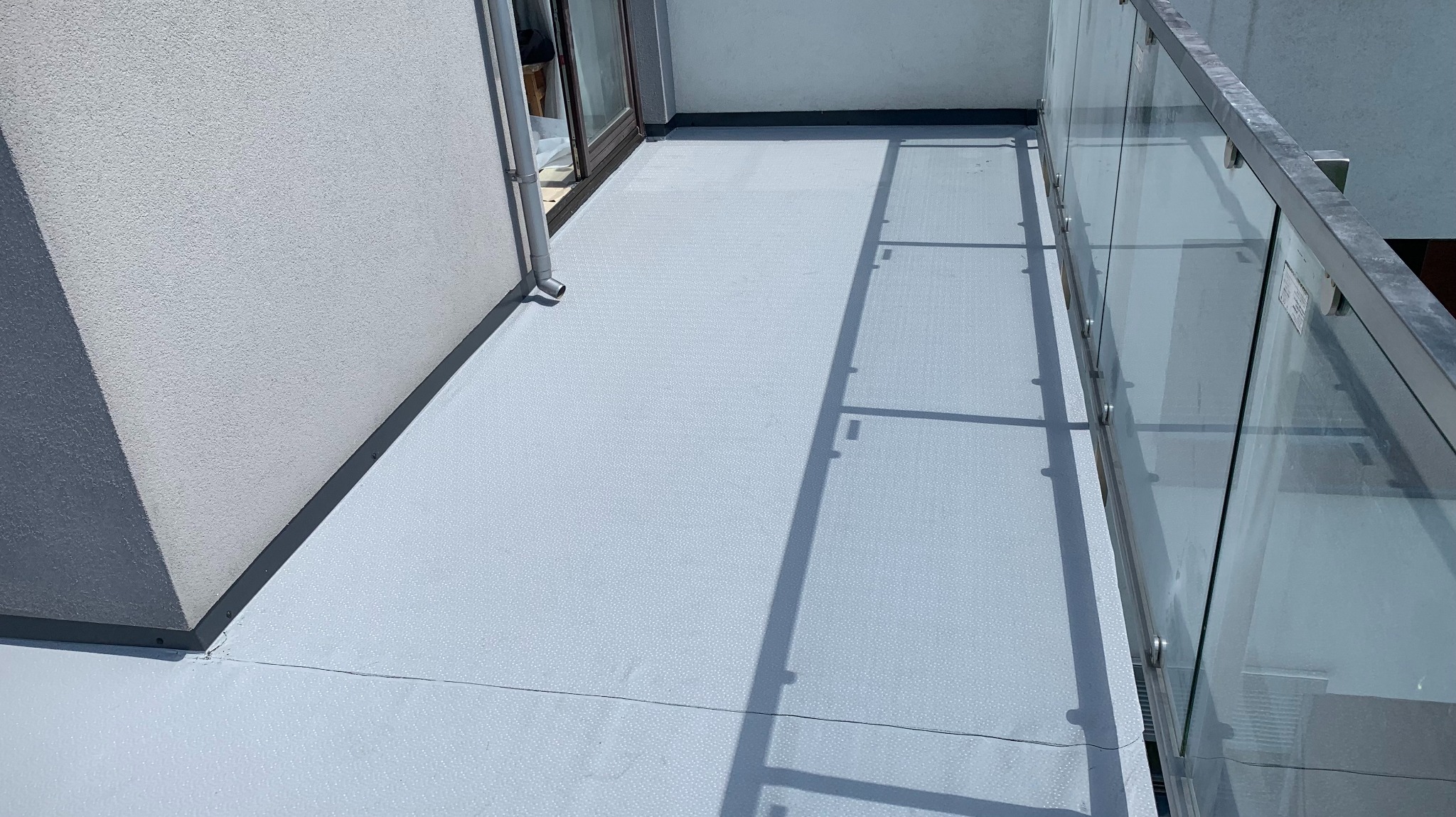 Wykończony balkon z szarą, teksturowaną podłogą, szklaną balustradą i odpływem rynnowym. Cień balustrady na podłodze. Widok z góry, jasne światło.