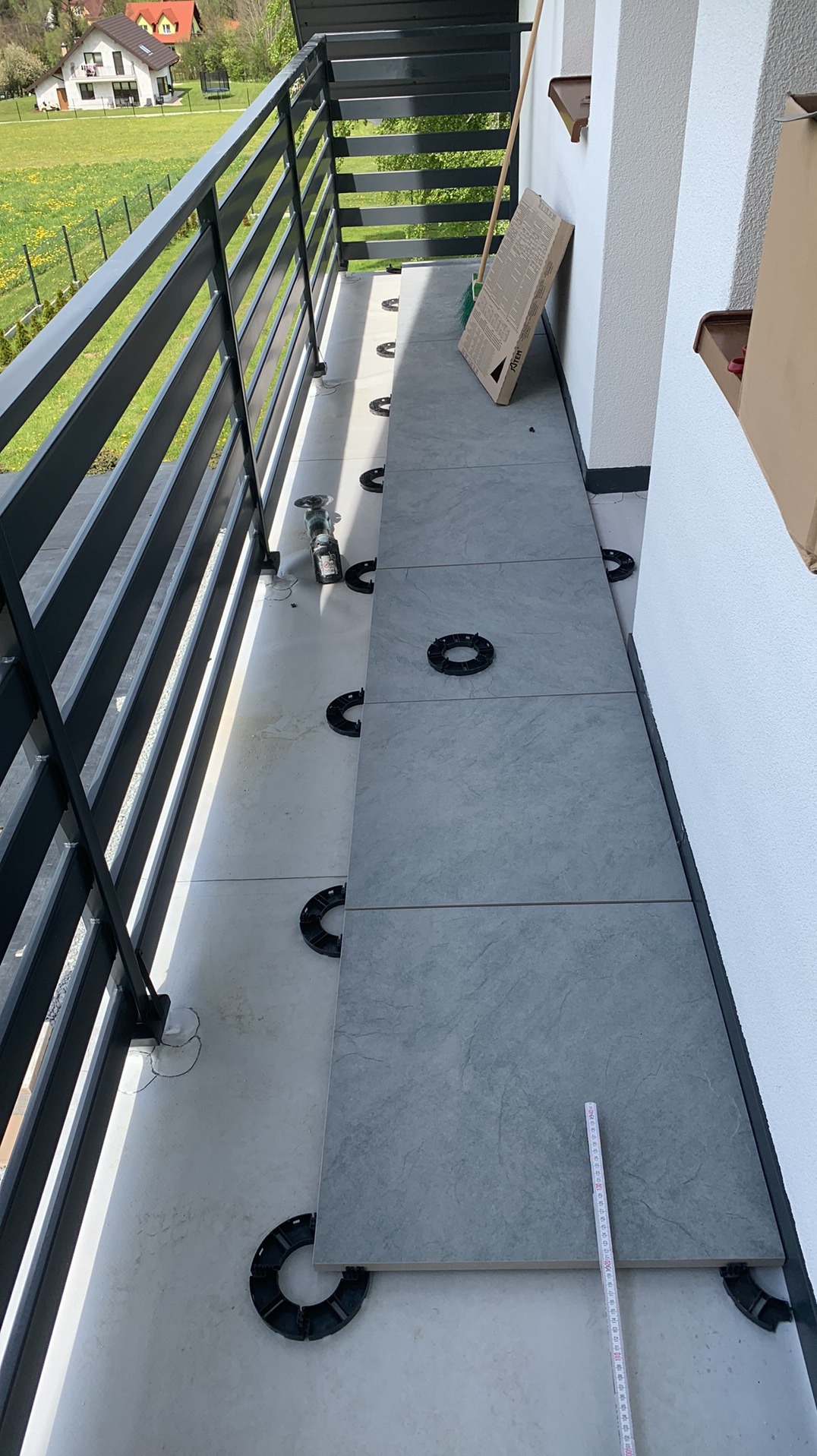 Montaż szarych płyt na wspornikach na balkonie z czarną balustradą. Widoczny fragment narzędzia i miarka. Prace w toku, częściowo ułożona podłoga.