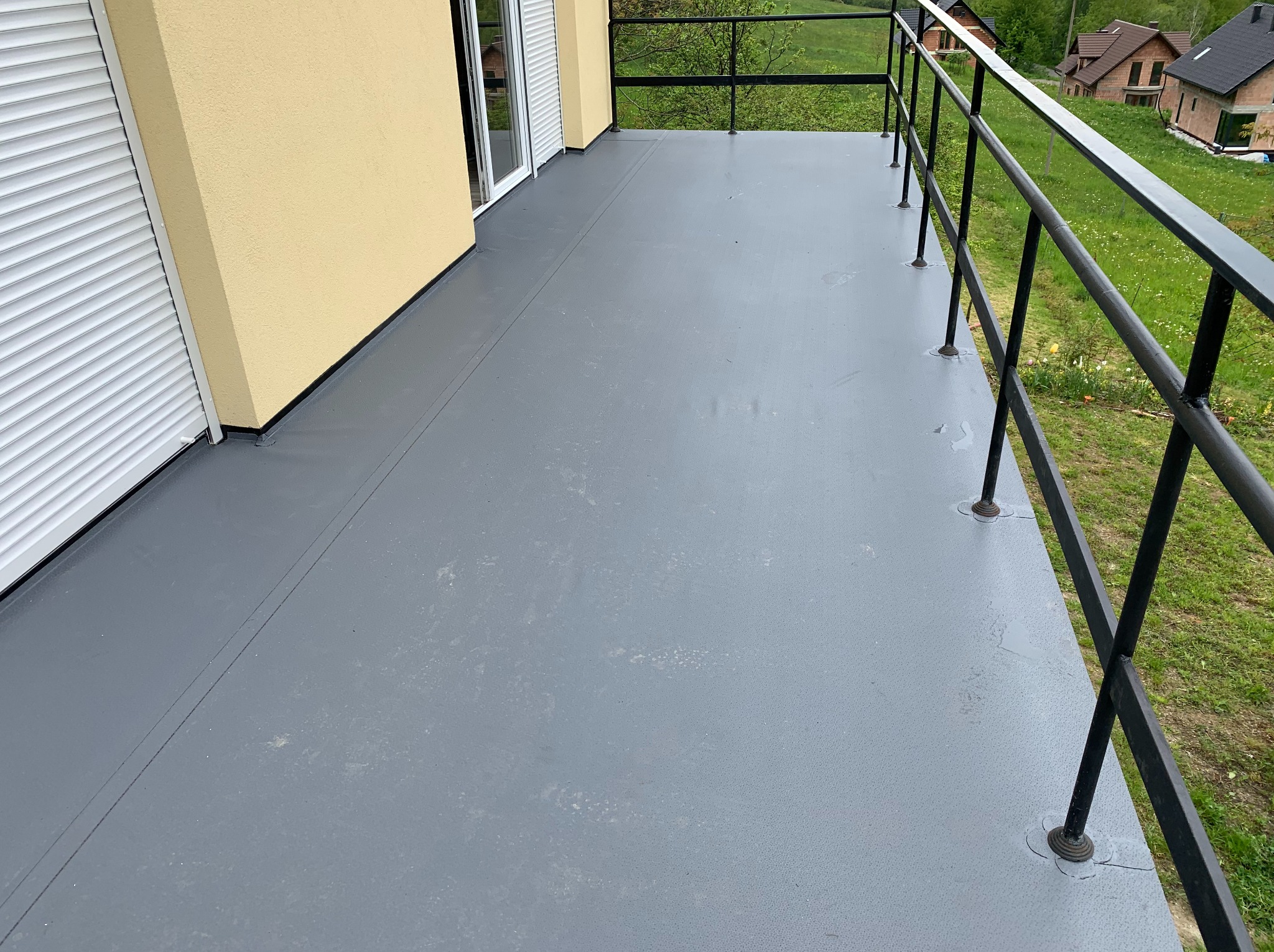 Szary, wykończony balkon z czarną metalową balustradą i żółtą ścianą budynku w tle. Widoczne rolety zewnętrzne i zieleń za balustradą.