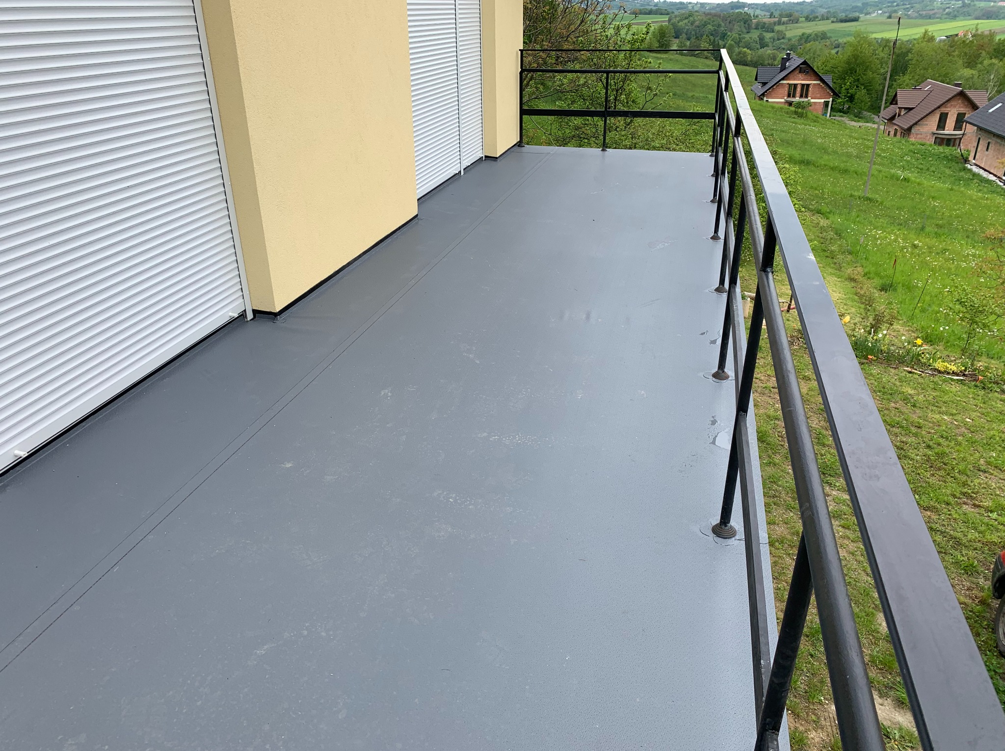 Szary, wykończony balkon z czarną balustradą i widokiem na zielone wzgórza. Żaluzje zewnętrzne i żółta elewacja budynku w tle. Nowoczesne wykończenie balkonu.