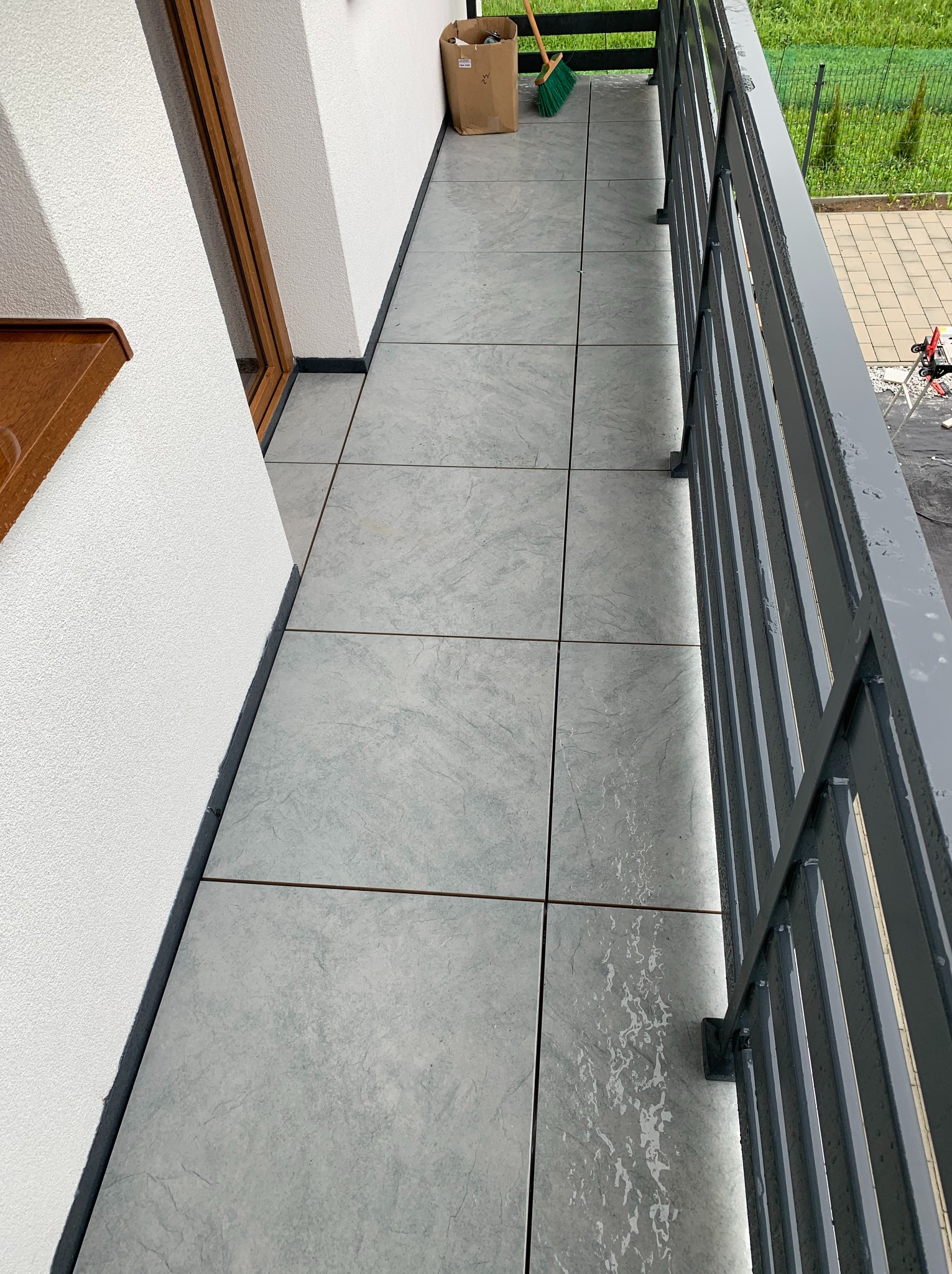 Wyłożony szarymi płytkami balkon z metalową balustradą. Widoczny fragment elewacji budynku i narzędzia pracy w tle. Świeżo położona podłoga.