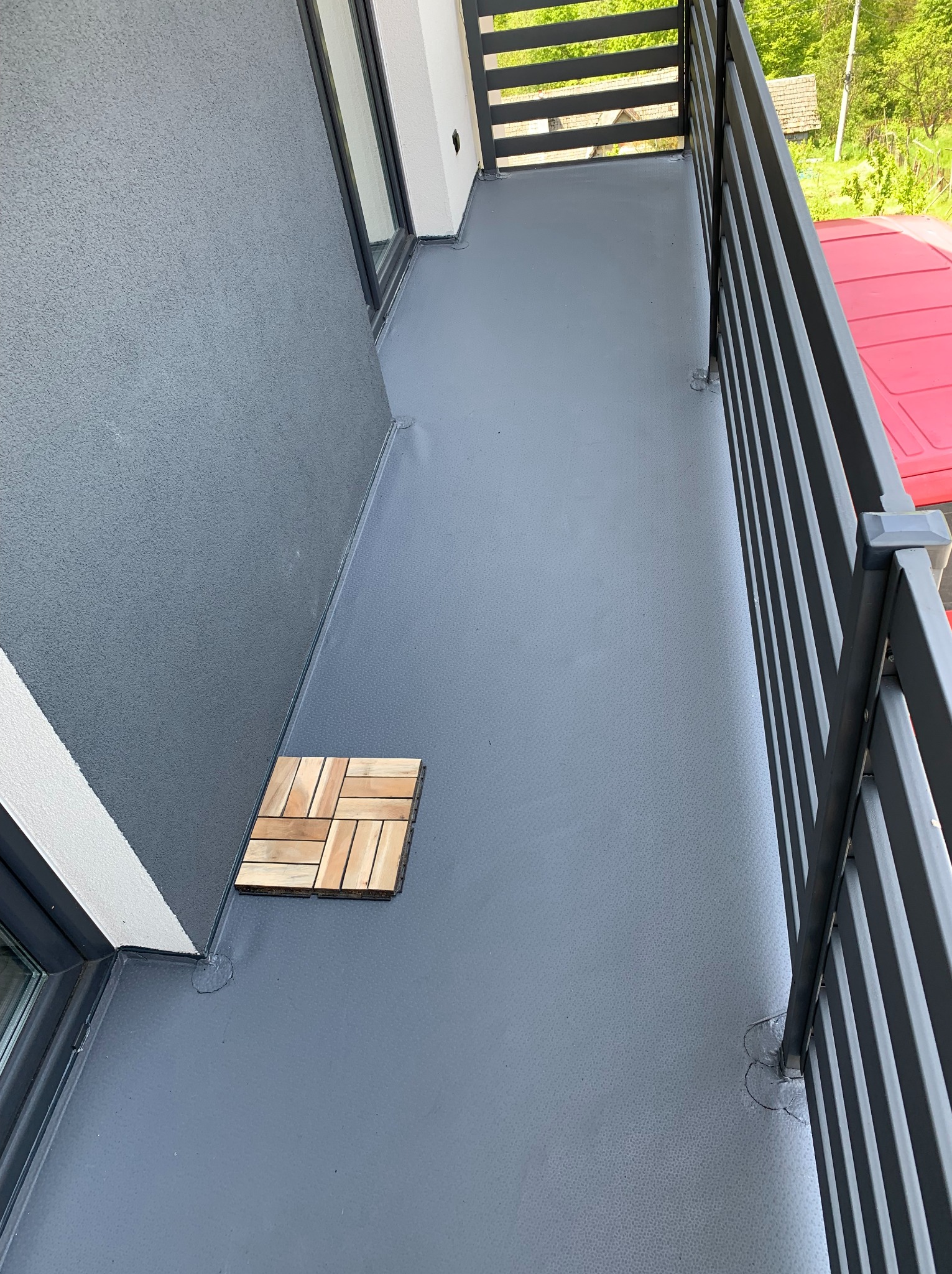 Szary balkon z drewnianymi płytkami podłogowymi, widok z góry. Nowoczesna balustrada i gładka powierzchnia podłogi. Detal uszczelnienia przy oknie.