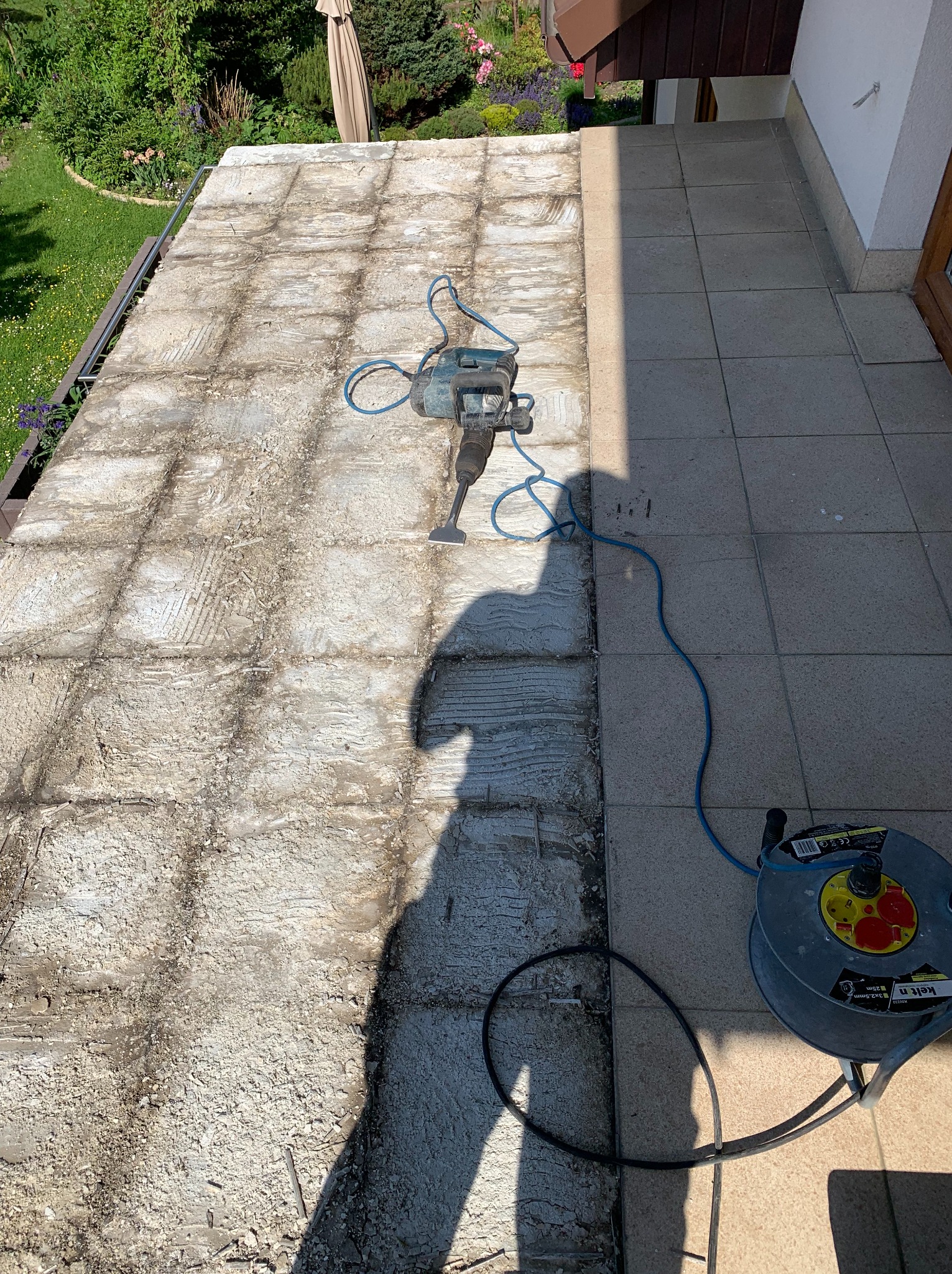 Demontaż starej posadzki na balkonie za pomocą młota udarowego. Częściowo usunięte płytki, widoczny kabel zasilający i cień osoby wykonującej prace.