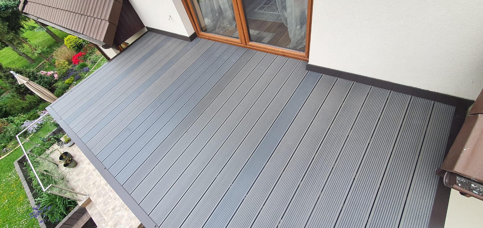 Nowoczesny balkon z szarą deską kompozytową, widok z góry. Balustrada ze szkła i stali nierdzewnej. Widok na ogród z parasolem. Estetyczne wykończenie.
