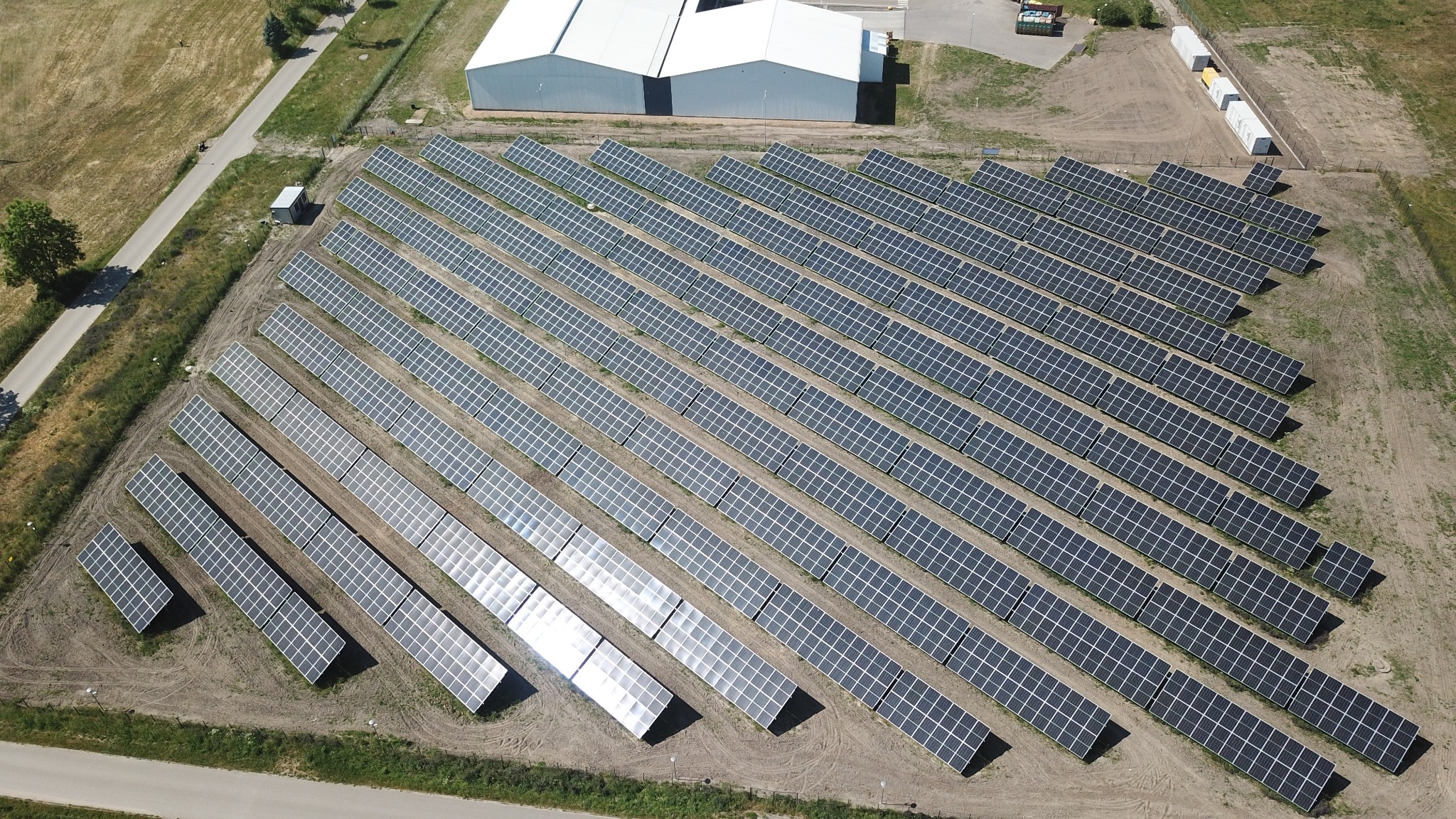 Widok z lotu ptaka na dużą farmę paneli słonecznych z budynkiem magazynowym w tle i drogą po lewej stronie.