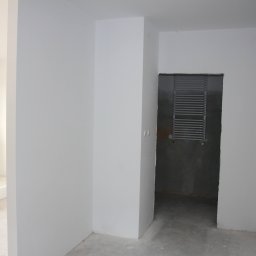 BARTŁOMIEJ ONDRUCH - Wnętrze w stanie deweloperskim: białe ściany, surowa podłoga, grzejnik drabinkowy w łazience. Widoczny domofon i fragment osoby po lewej stronie kadru.