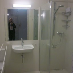 BARTŁOMIEJ ONDRUCH - Jasna łazienka z kabiną prysznicową i umywalką. W lustrze odbija się osoba robiąca zdjęcie. Minimalistyczny design, białe płytki podłogowe, proste oświetlenie.