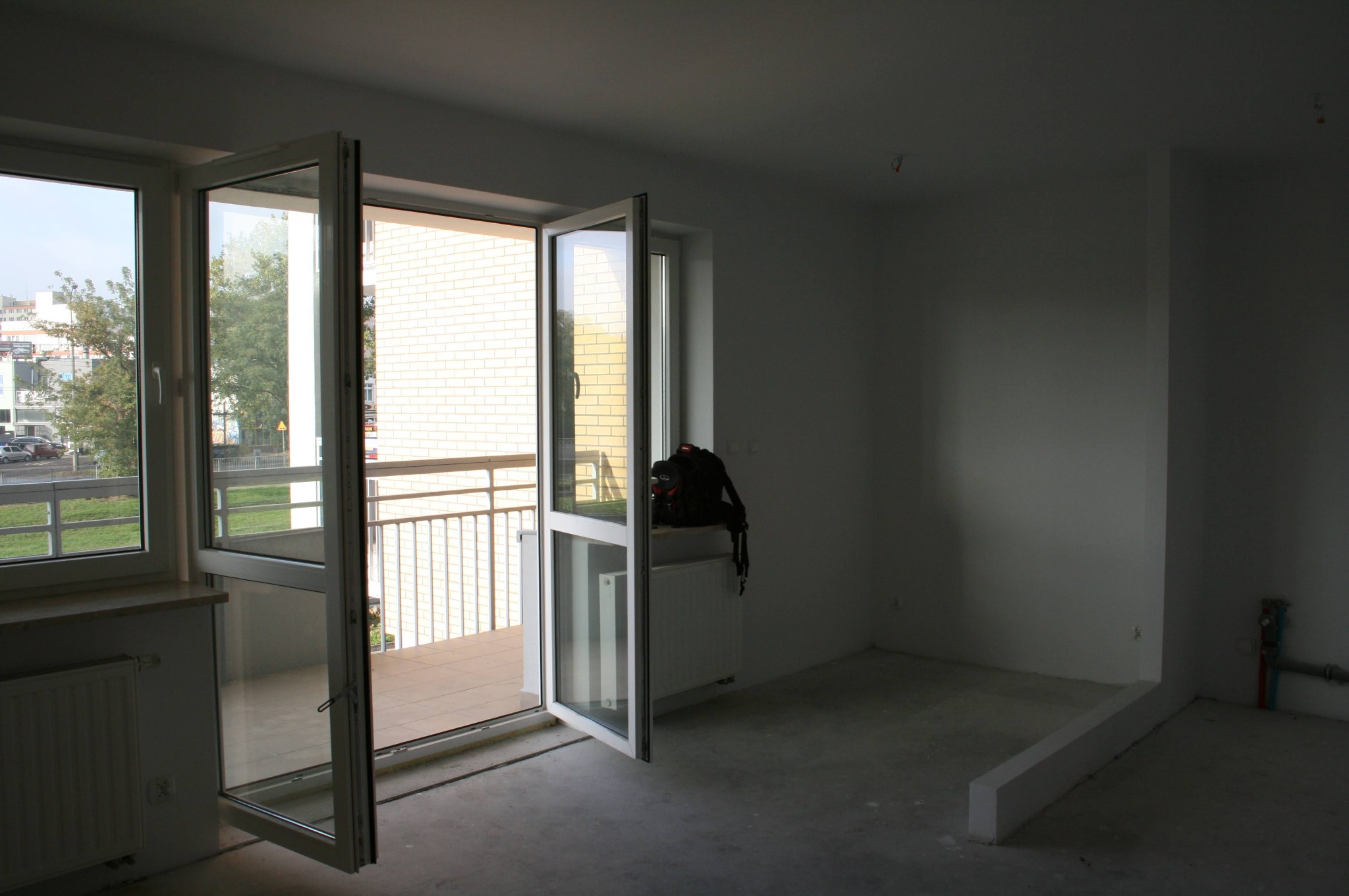 Surowe wnętrze mieszkania w stanie deweloperskim z otwartymi drzwiami balkonowymi. Widok na balkon i okolicę. Plecak leży na grzejniku.