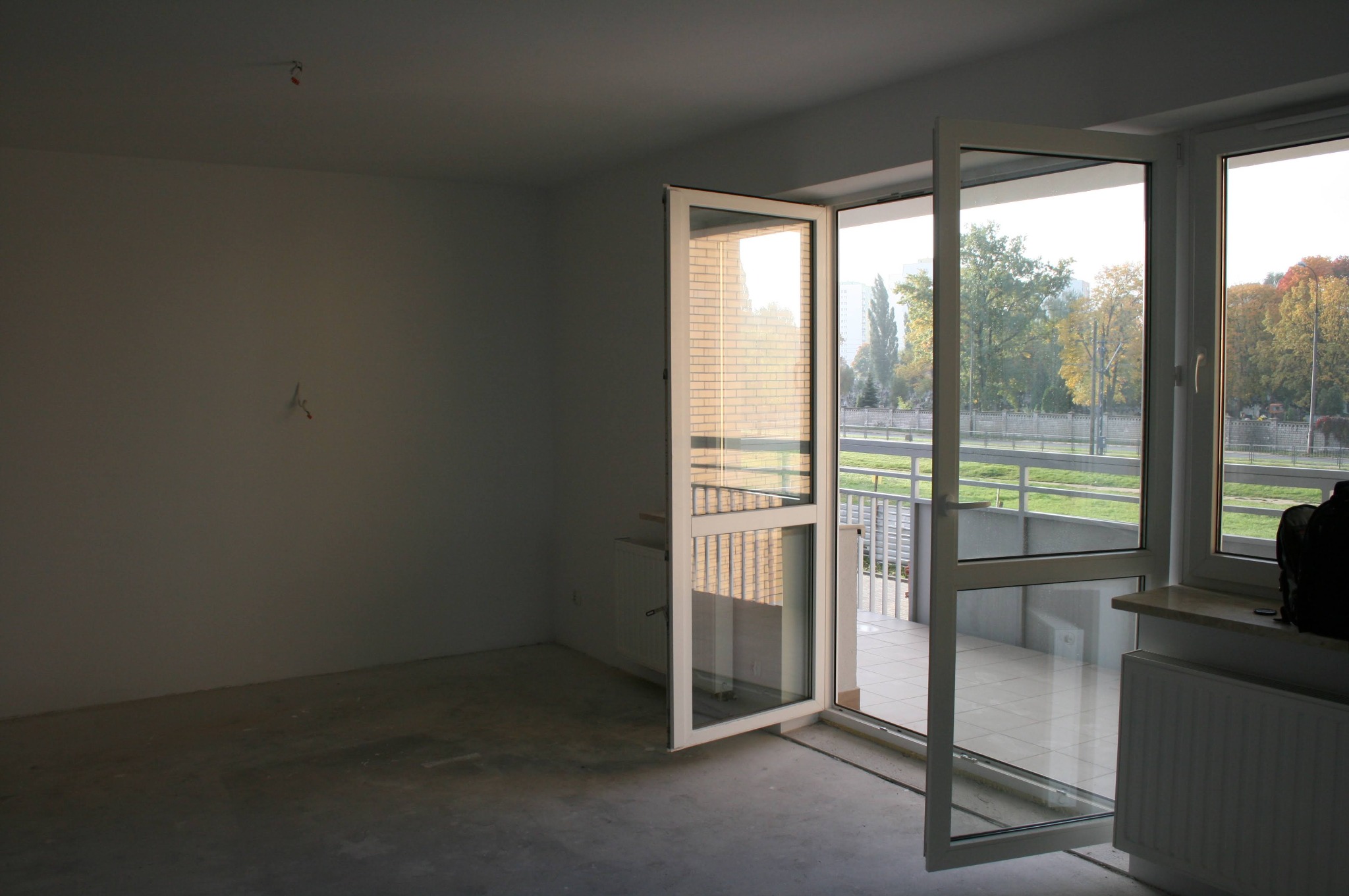 Pusty pokój w stanie deweloperskim z otwartymi drzwiami balkonowymi, widok na zewnątrz. Minimalistyczne wnętrze z surowymi ścianami i betonową podłogą.